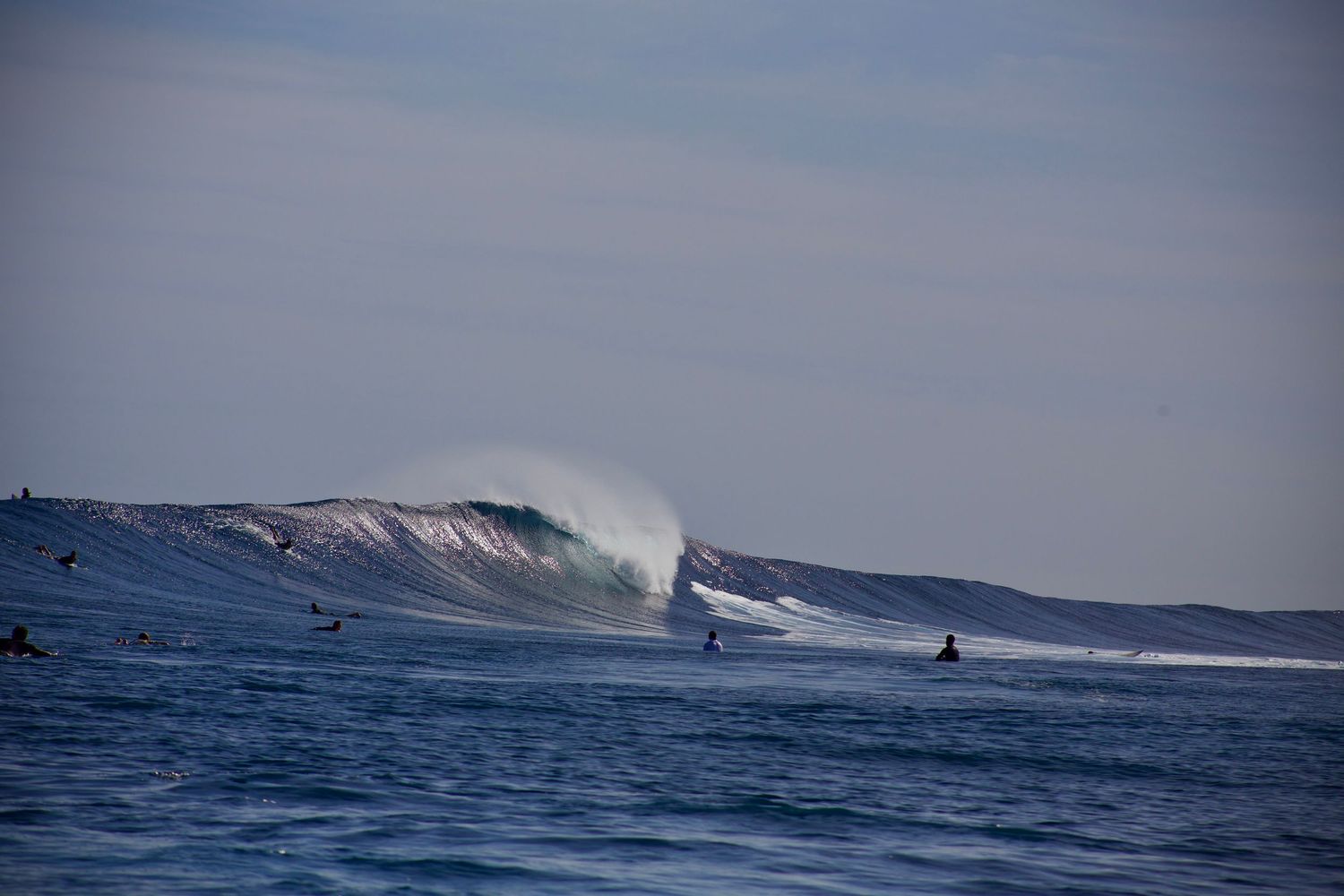 Surfer rides massive ocean wave with white spray trailing behind as others wait in the distance on a cloudy day in the South Pacific in 2025