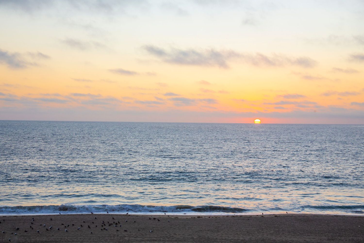 A tranquil ocean sunset with orange sky reflecting on calm waters as gentle waves roll onto a dark sandy beach.