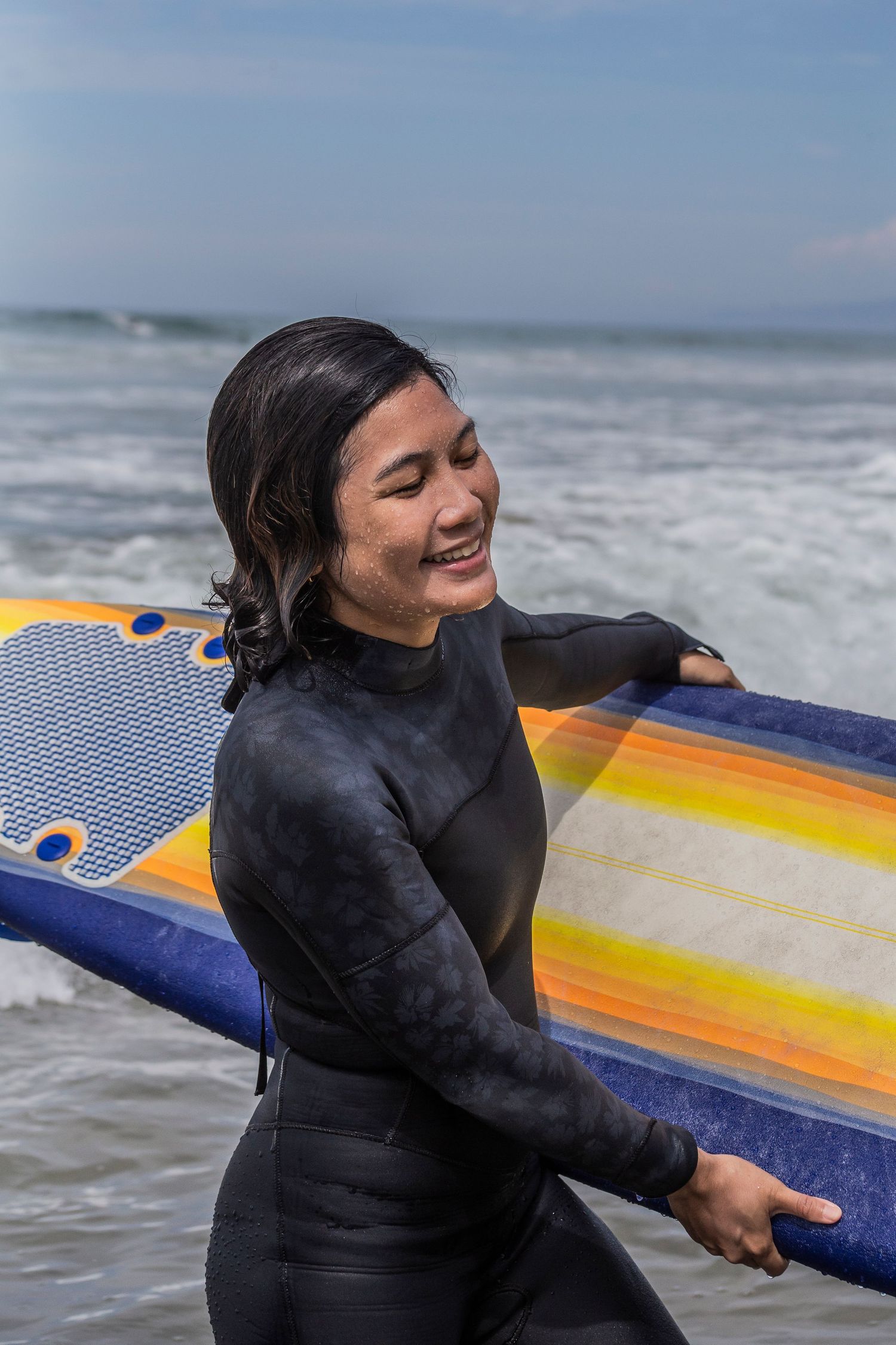 A beginner surfer in a wetsuit smiles joyfully while holding a surfboard at the beach with crashing waves in the background.
