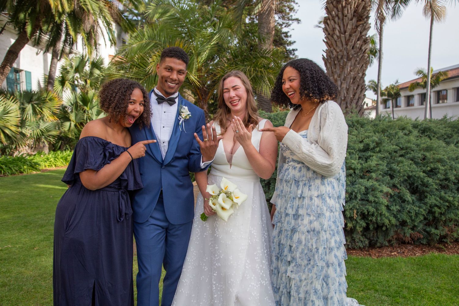 Wedding party poses together outdoors with palm trees and lush greenery in the background.