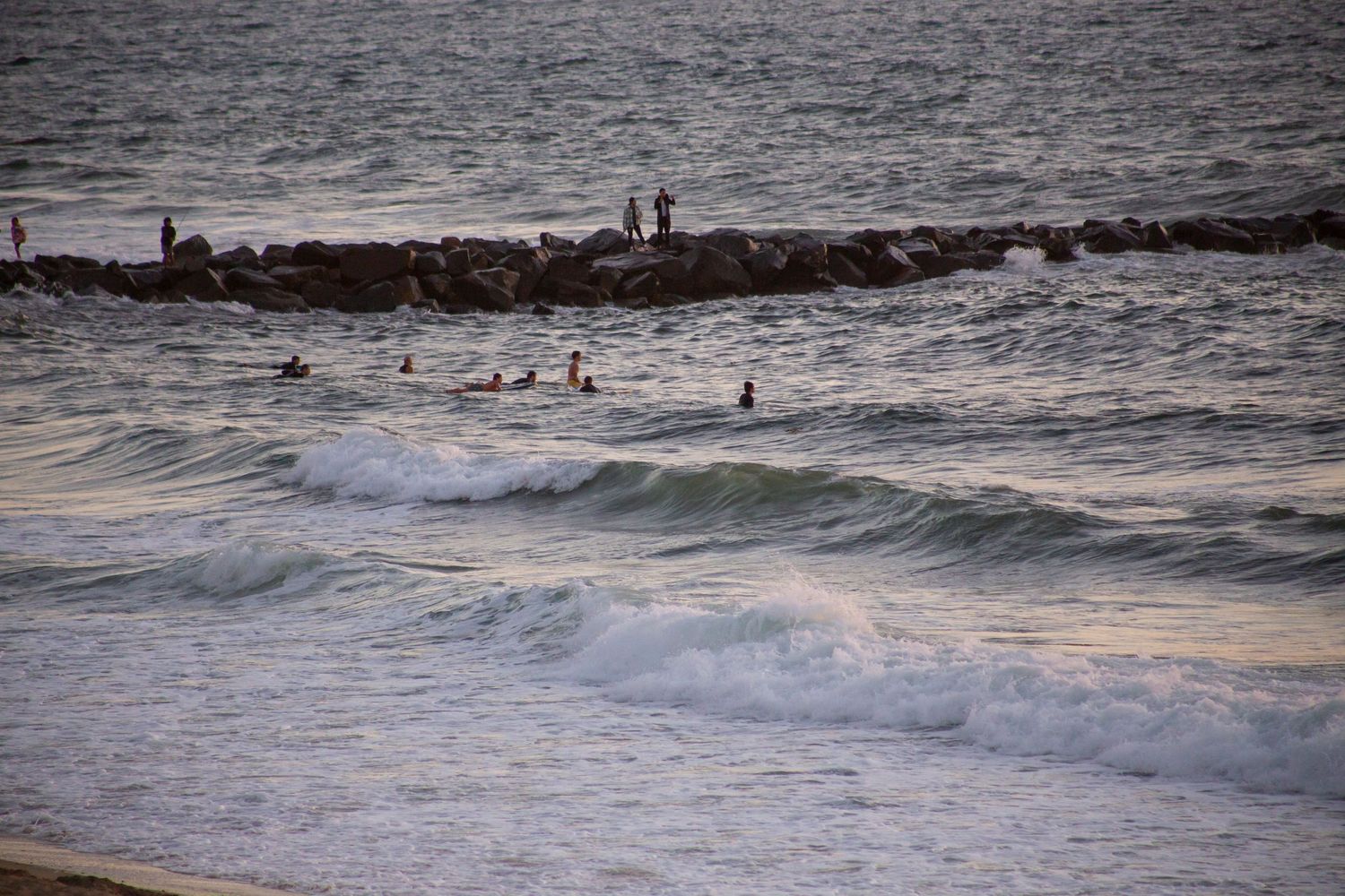 Aerial view of surfers riding waves at sunset along a rocky coastline.