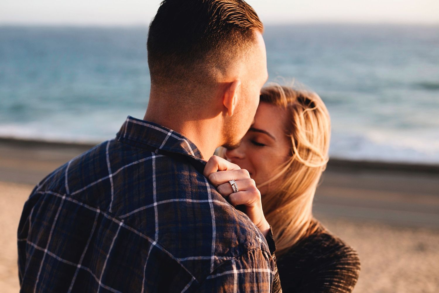 Romantic engaged couple sharing intimate moments at sunset on a sandy beach with ocean waves in background.