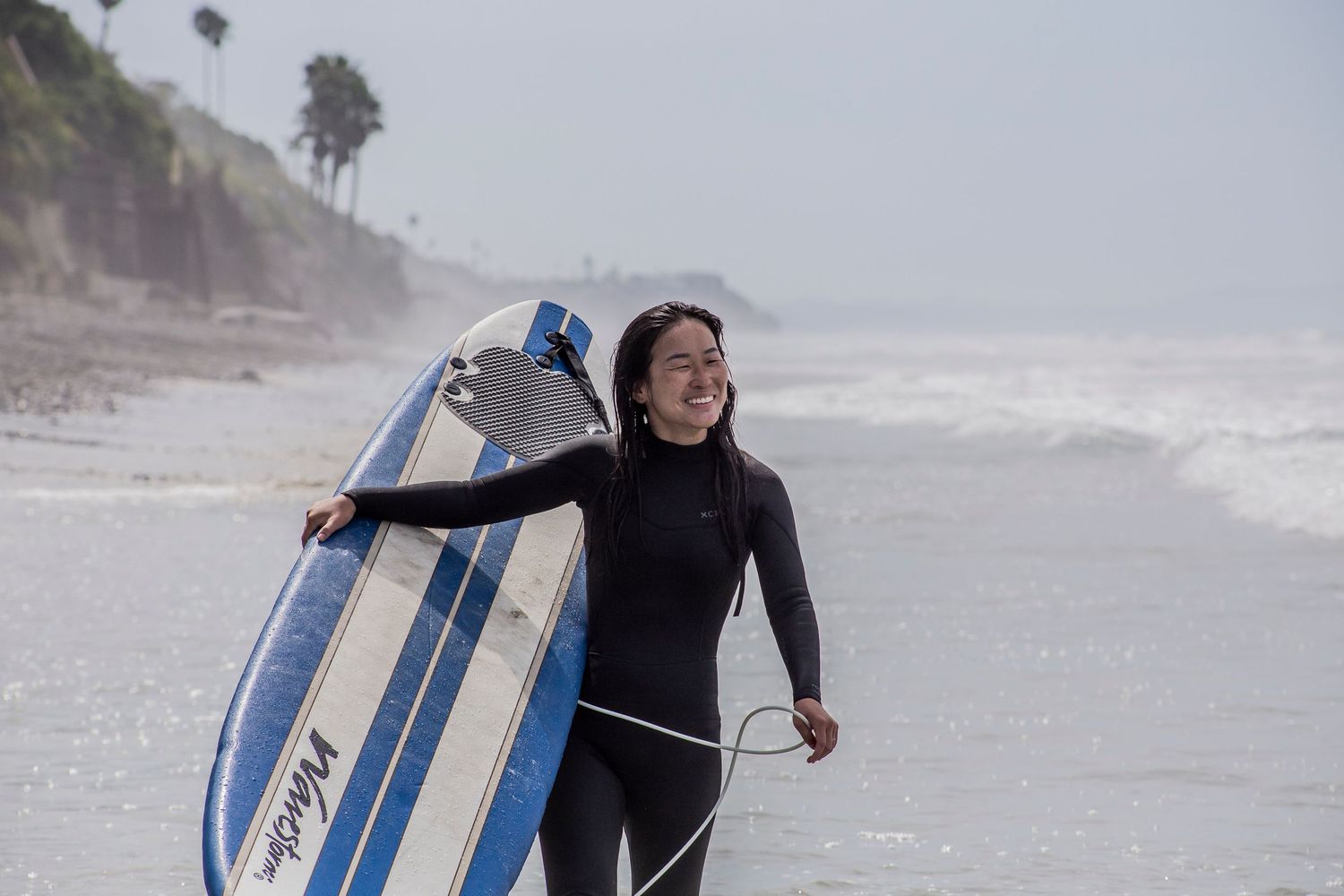 A young female surfer in a black wetsuit carries a blue and white surfboard along a misty beach shoreline.