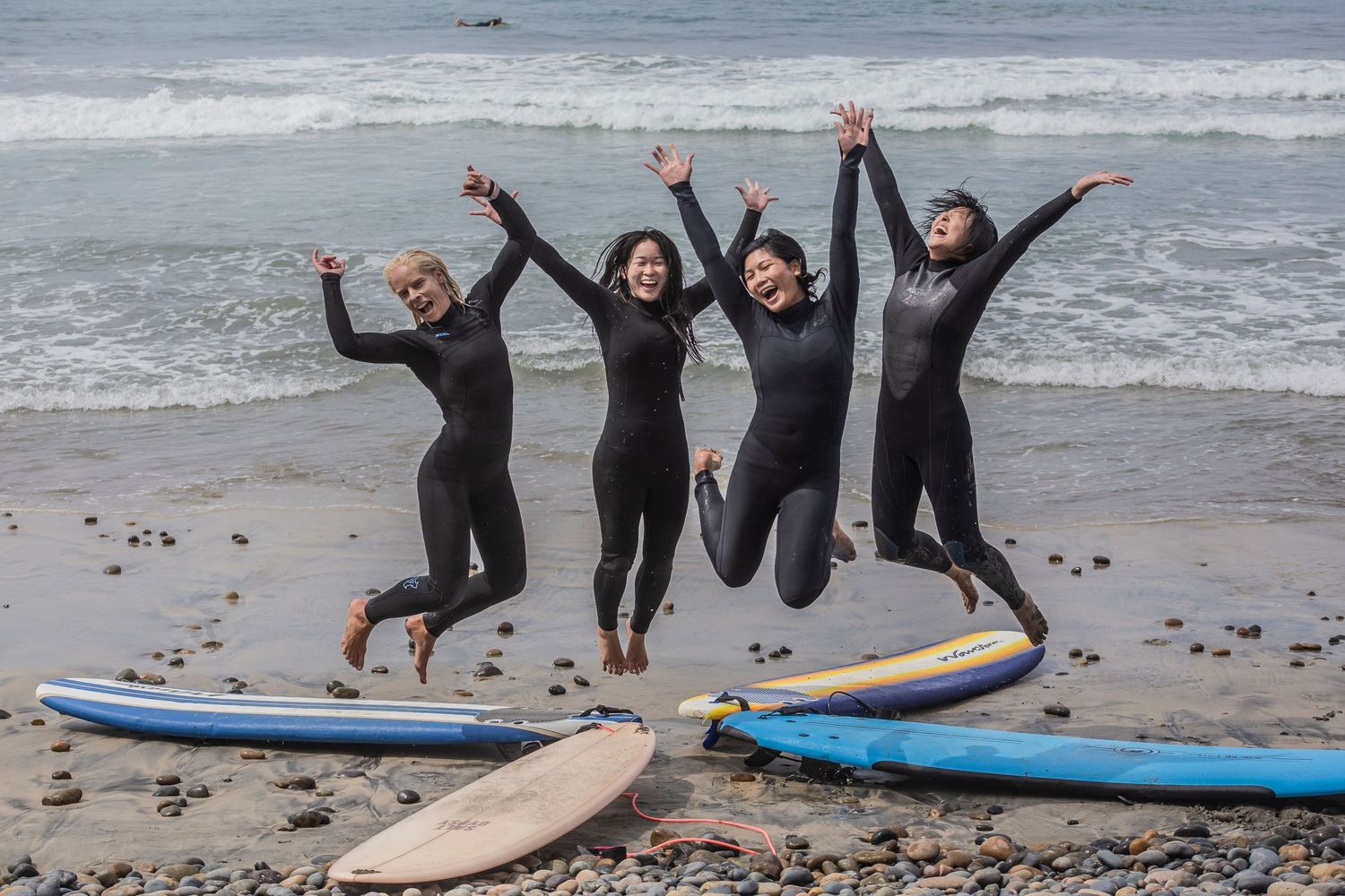 A group of surfers in wetsuits celebrate together on a rocky beach with surfboards nearby.