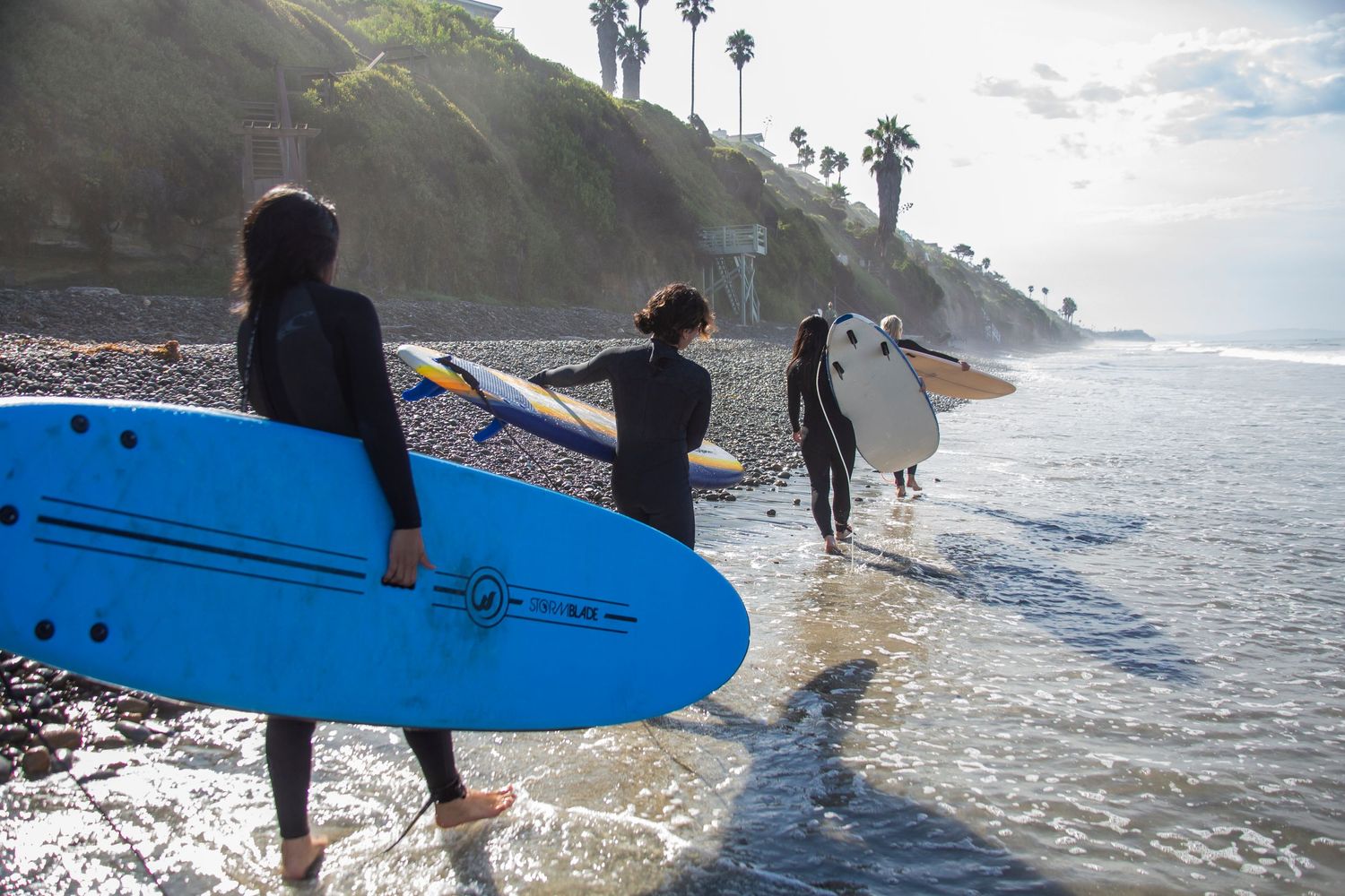 Surfers carrying surfboards walk along a scenic beach with palm trees and cliffs during mid-day in San Diego.