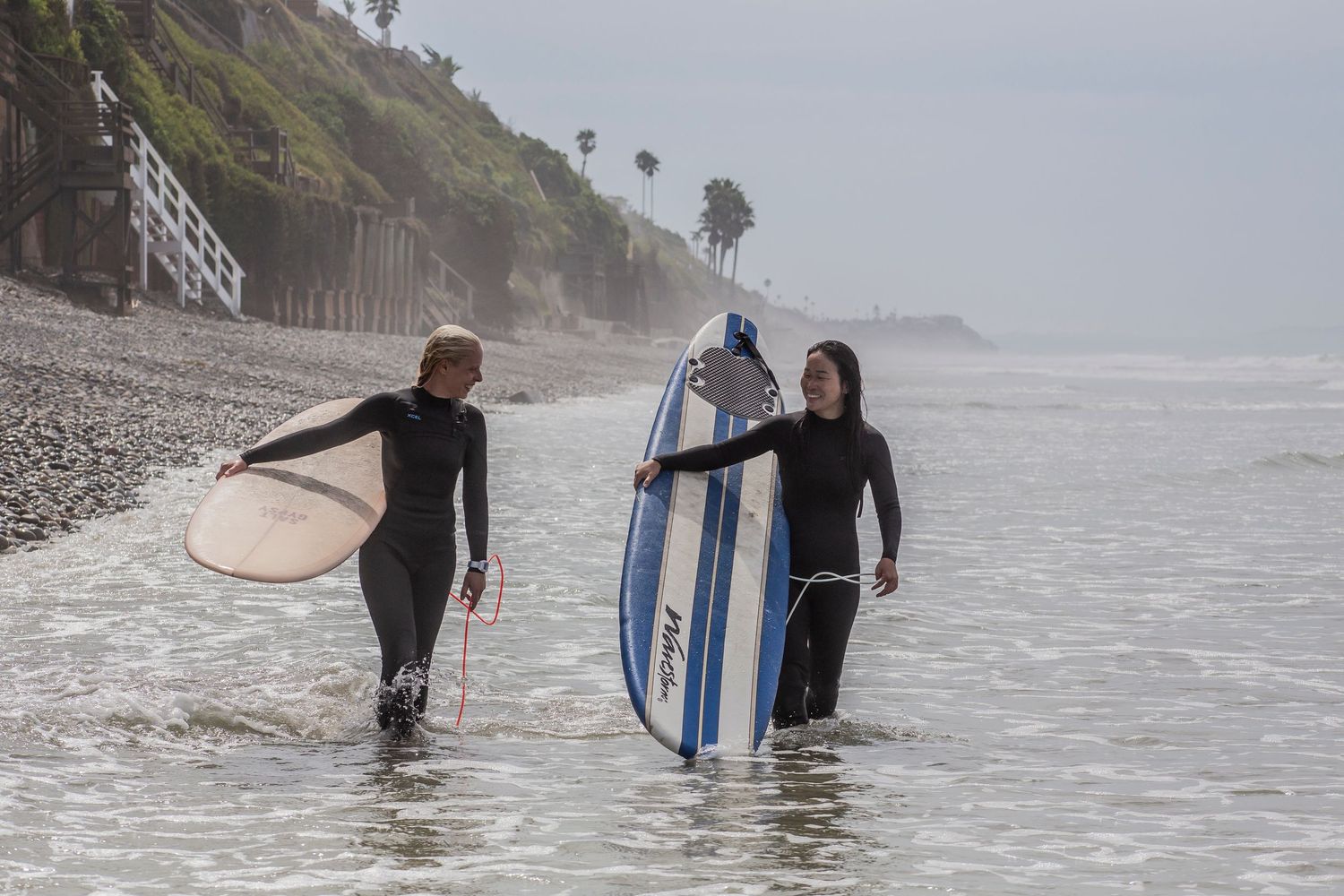 Surfers carrying their boards into the misty ocean along a scenic coastal beach on a foggy day.