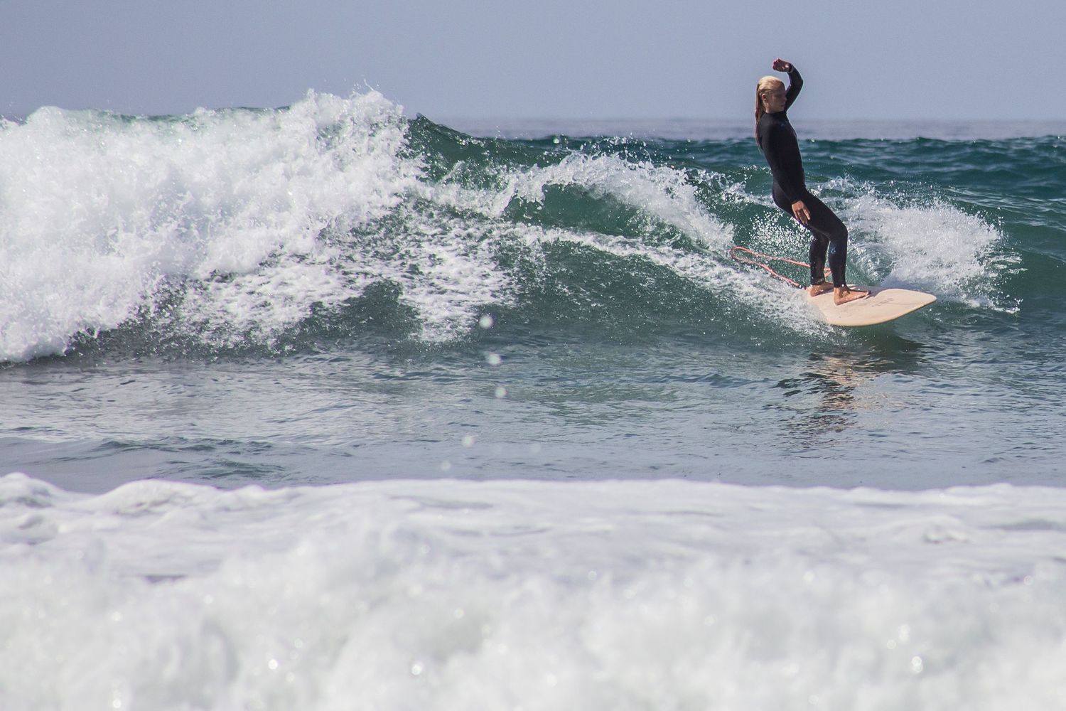 A female surfer rides a wave on a surfboard while ocean spray creates a dramatic backdrop in the blue-green waters.