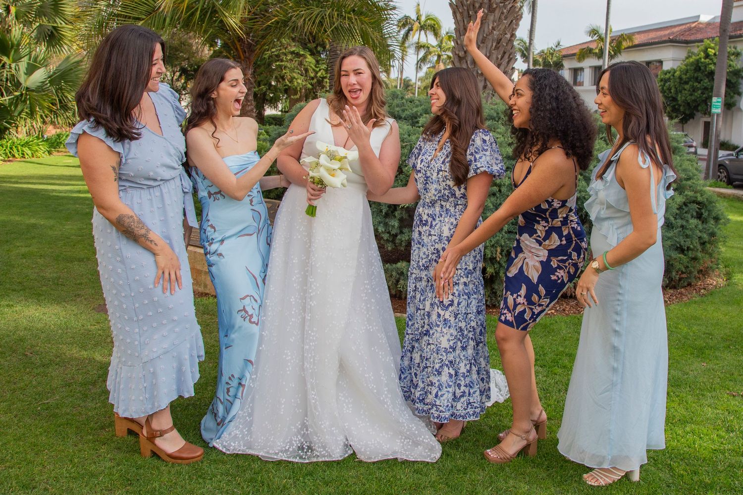 A group of bridesmaids and bride in pastel dresses laugh together on a grassy lawn during a sunny outdoor wedding celebration.