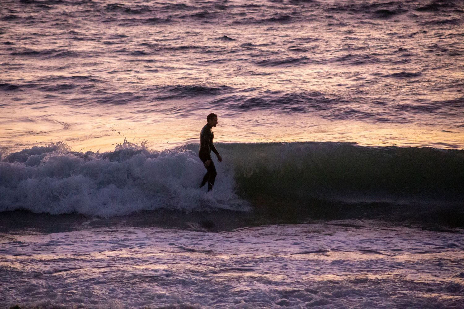 A surfer rides waves during a vibrant purple and orange sunset at the beach.