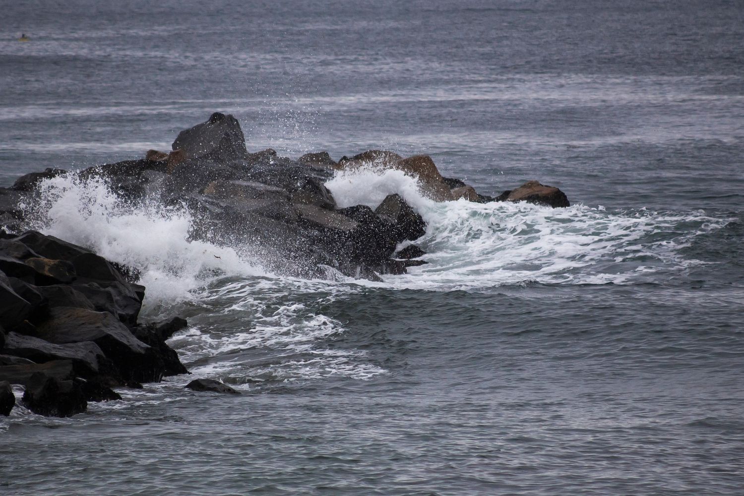 Dramatic ocean waves crash against dark rocky cliffs along a rugged coastline during stormy weather.