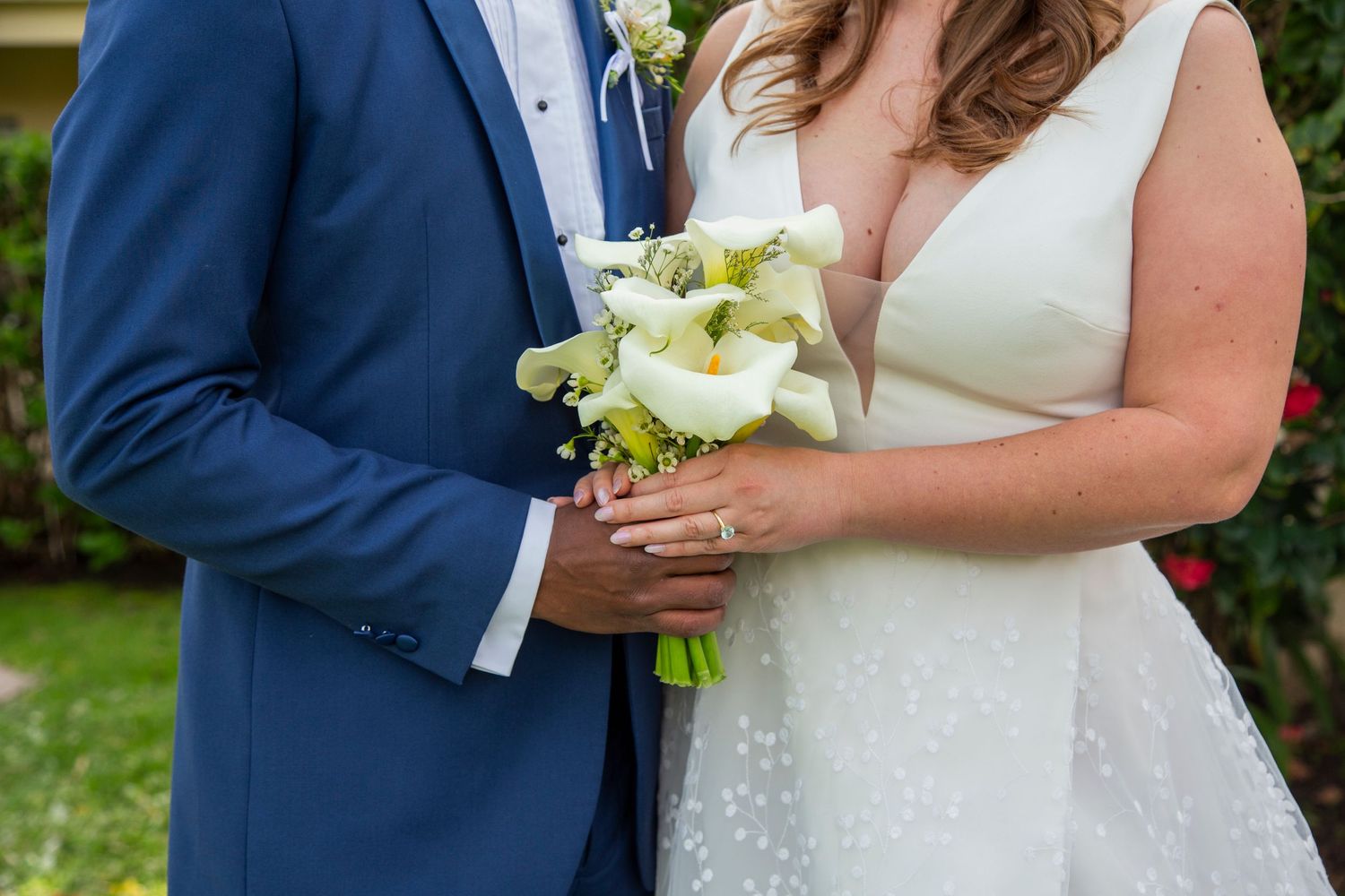 Wedding couple holds hands and embraces while standing in a lush garden setting.