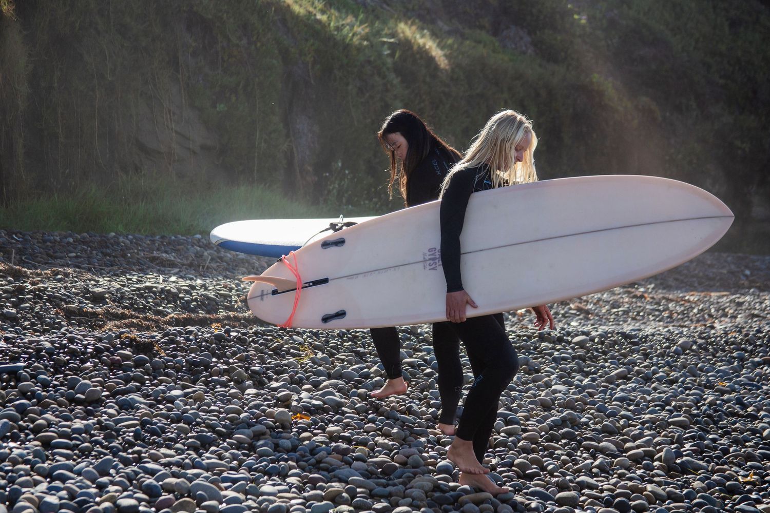 Female empowerment surfers walking on a rocky beach carrying surfboards during an overcast day.