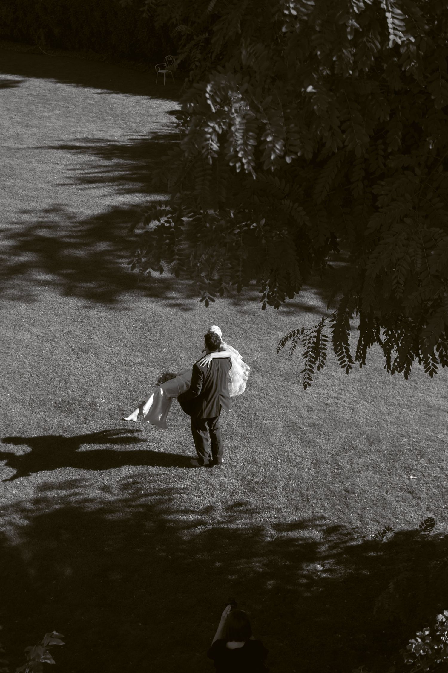 Black and white aerial view of a person walking across a sunlit lawn carrying a large white bag near trees.