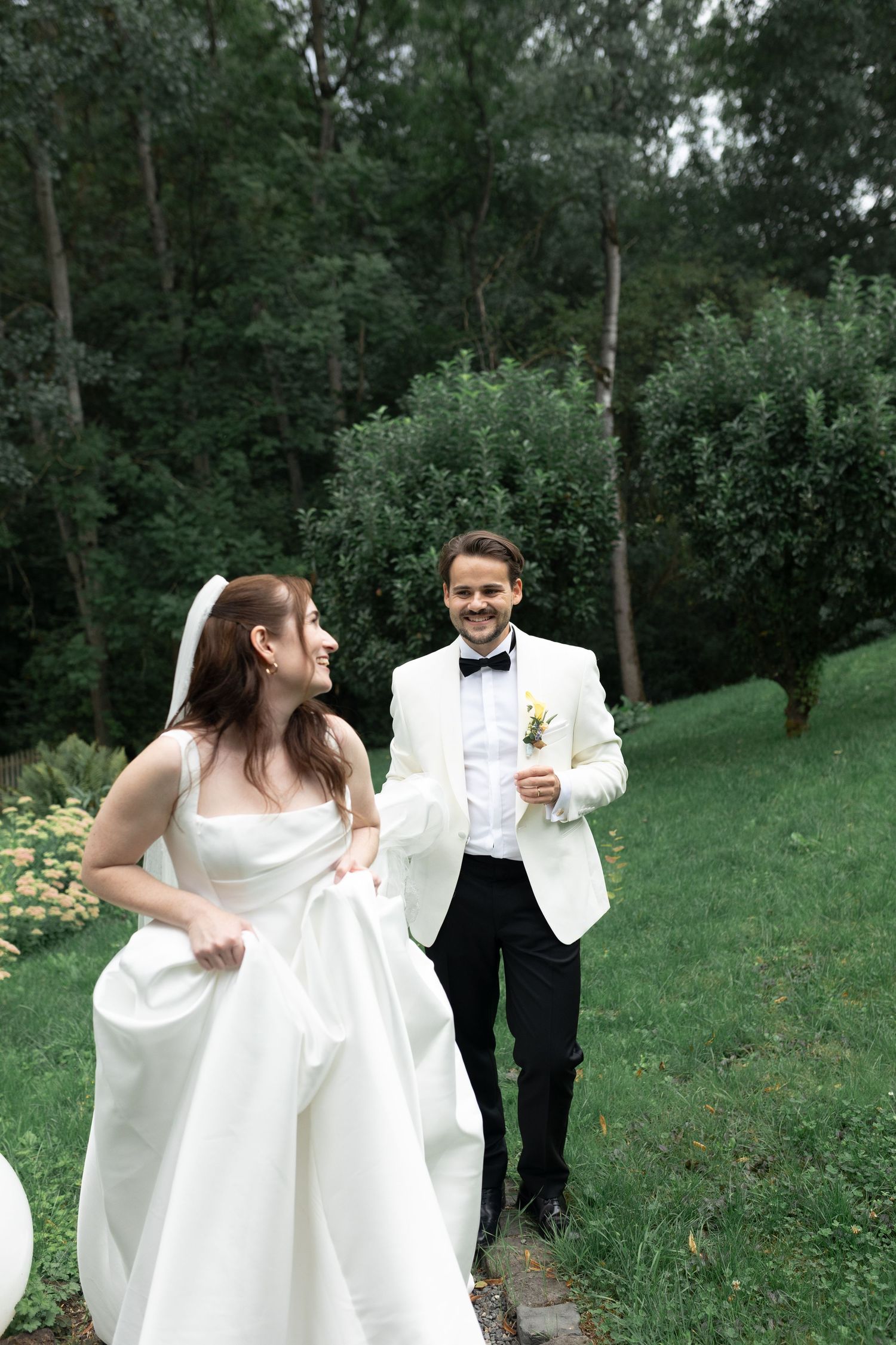 Happy bride and groom walking outdoors in lush green garden, wearing white wedding dress and white tuxedo.