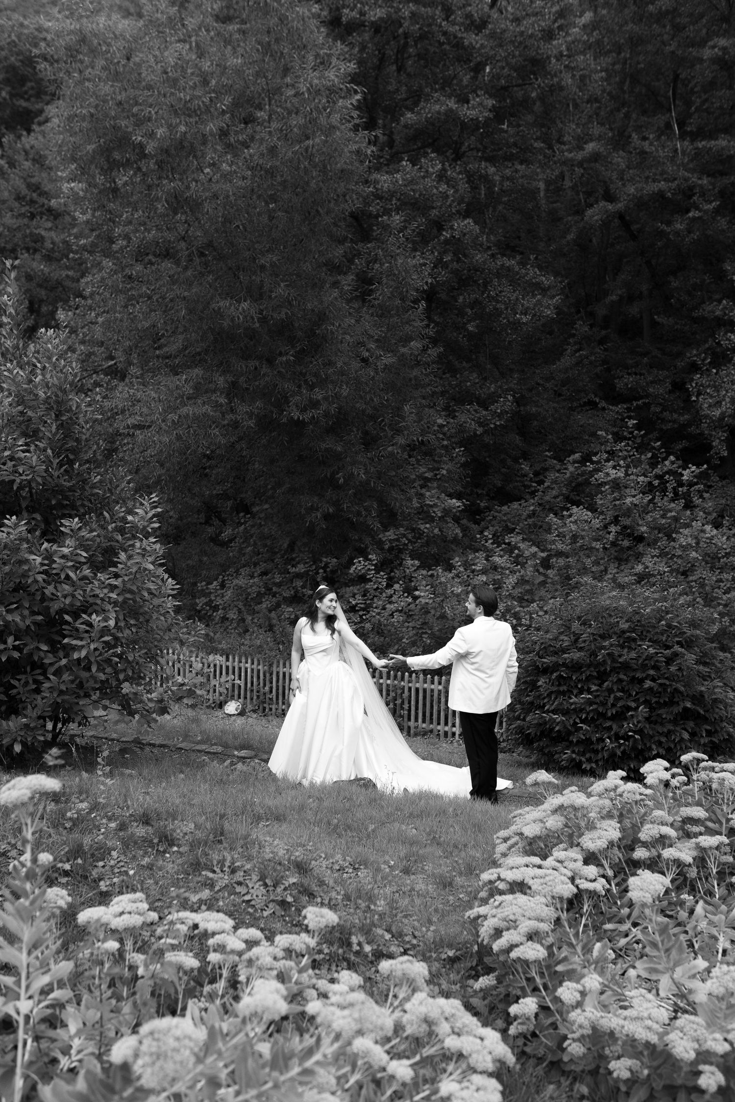Black and white photo of a bride and groom holding hands in a lush garden surrounded by wildflowers and dense trees.