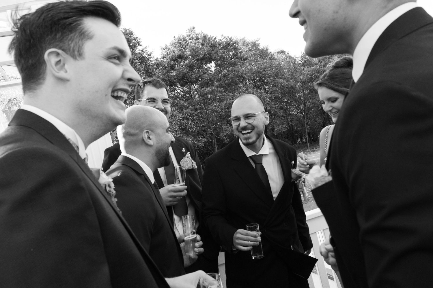 Black and white photo of men in suits laughing and socializing at a formal outdoor wedding reception.