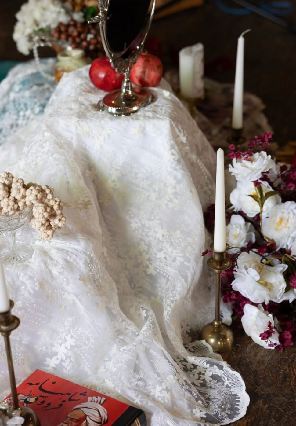 Elegant Persian Sofreh Aghd wedding spread with lace fabric, candles, pomegranates, mirror, and floral arrangements.