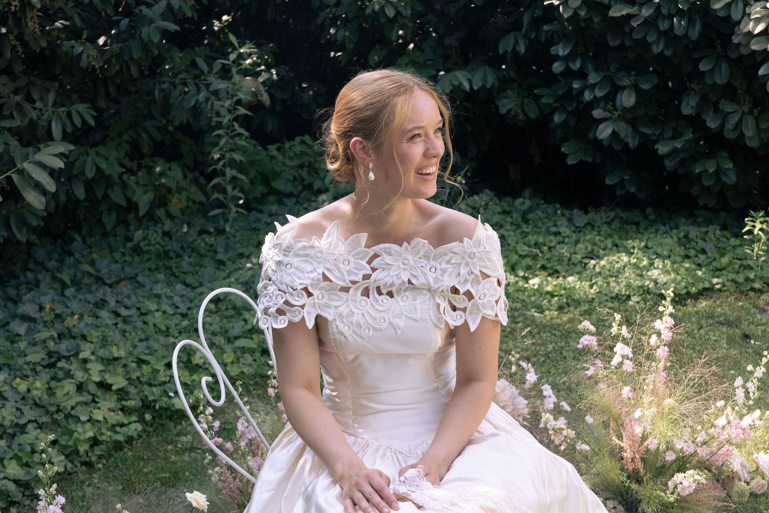 Bride in white lace off-shoulder gown sitting on garden bench surrounded by lush green foliage.