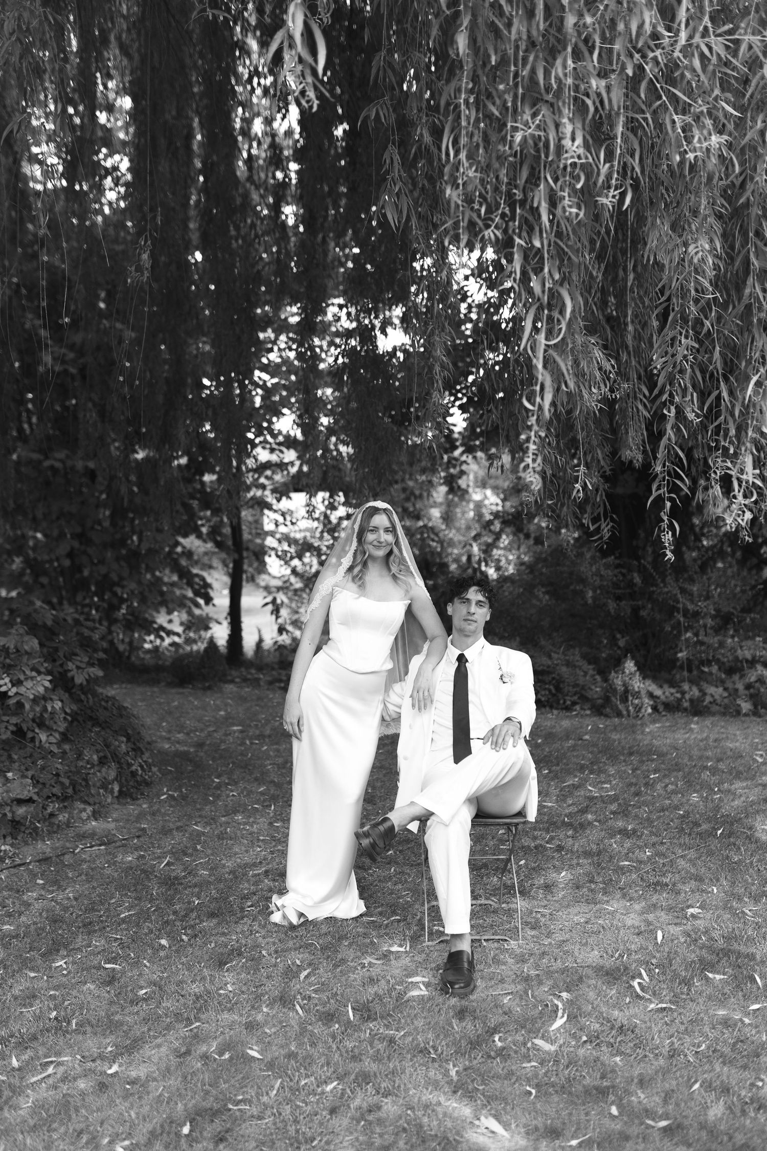 Black and white photo of a bride and groom posing under a weeping willow tree outdoors.