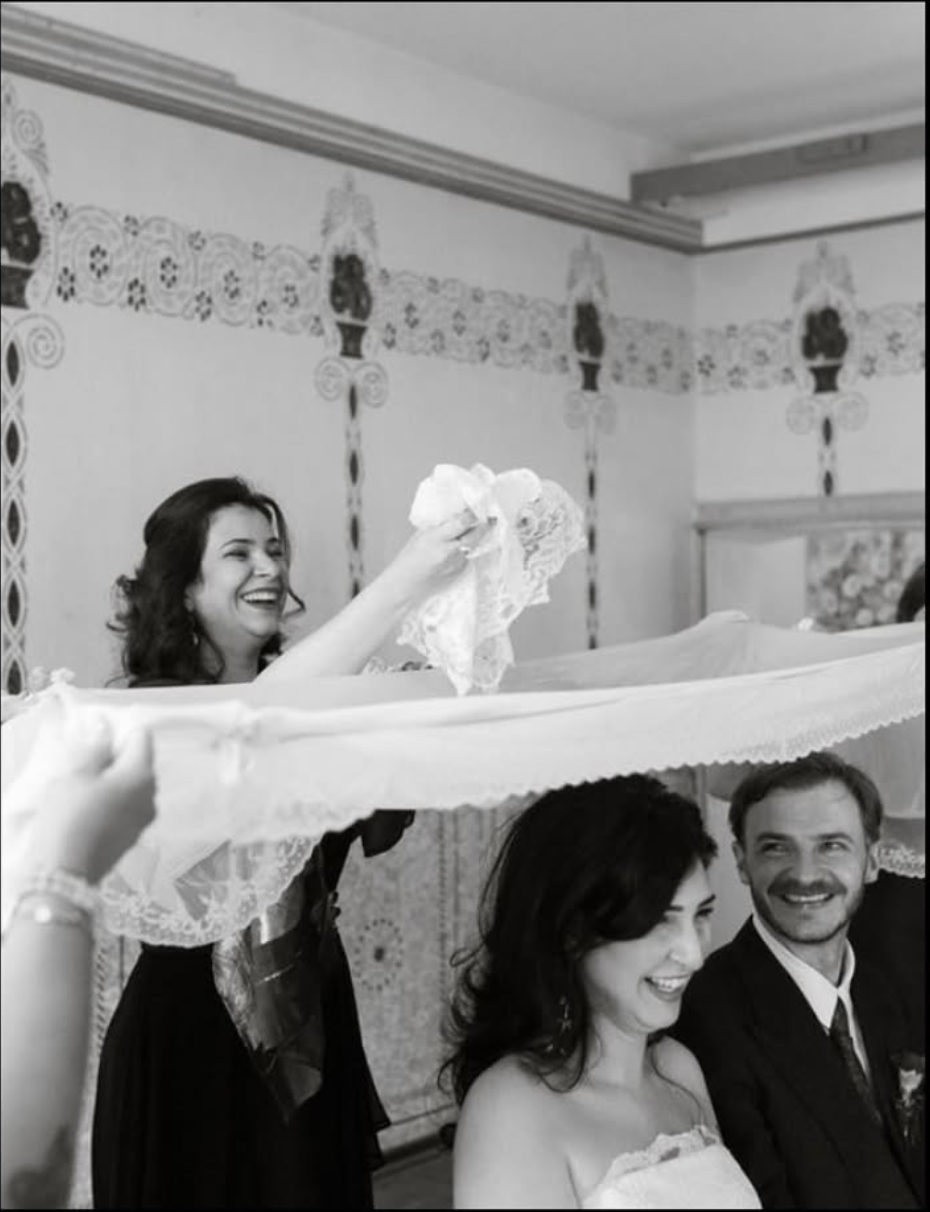 Black and white wedding photo of bride smiling as attendant lifts veil while groom watches joyfully.