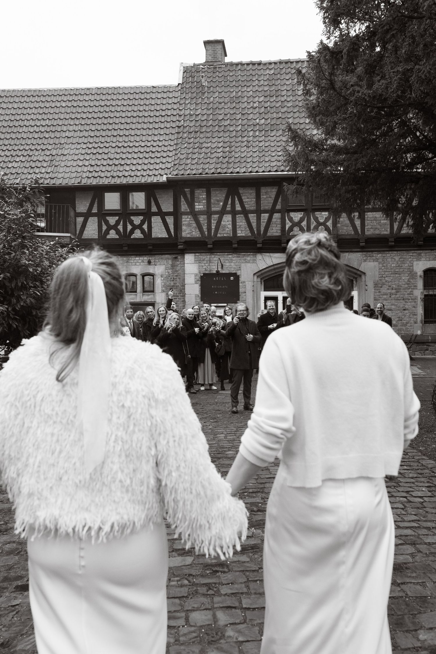 Two brides in white dresses hold hands walking toward wedding guests outside a historic half-timbered building.