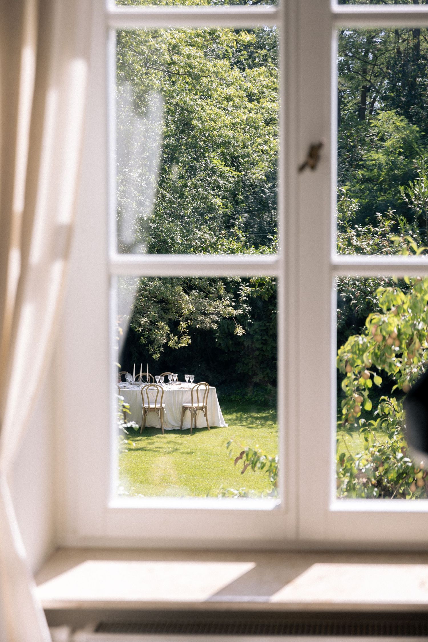 White-framed window overlooking an elegant outdoor garden dining table set on lush green lawn surrounded by trees.