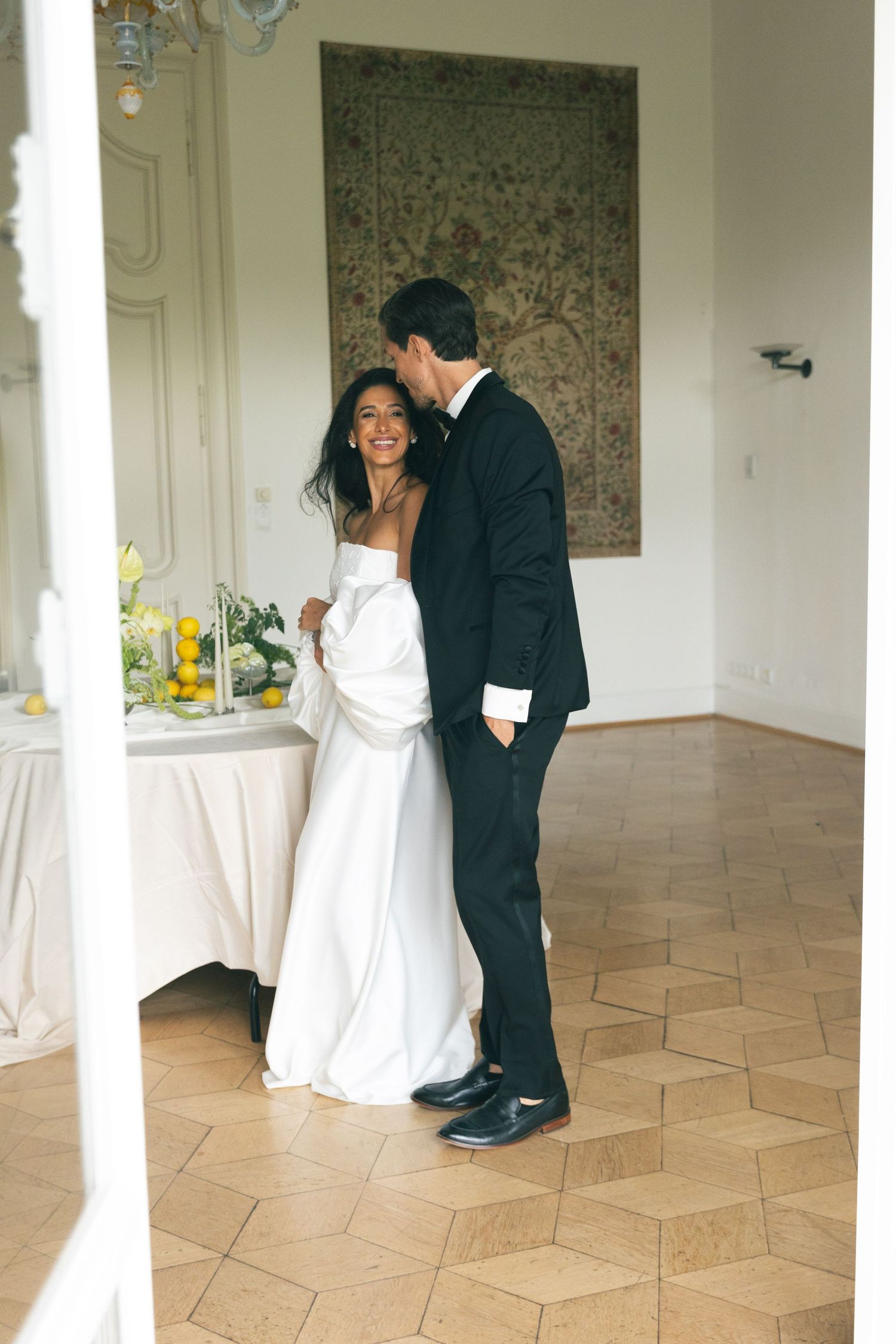 A smiling bride in a white gown stands with a groom in a black tuxedo in an elegant room with a tapestry.