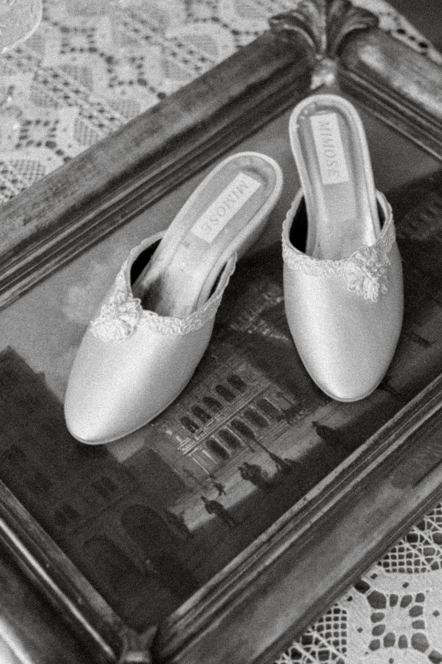 Elegant white satin bridal mules with lace detail displayed on a vintage framed painting, black and white photo.