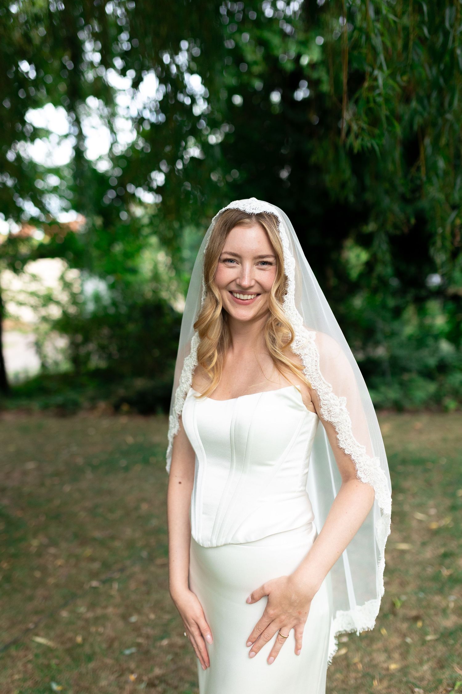 Smiling bride in white corset gown and lace veil posing outdoors in a lush green garden setting.
