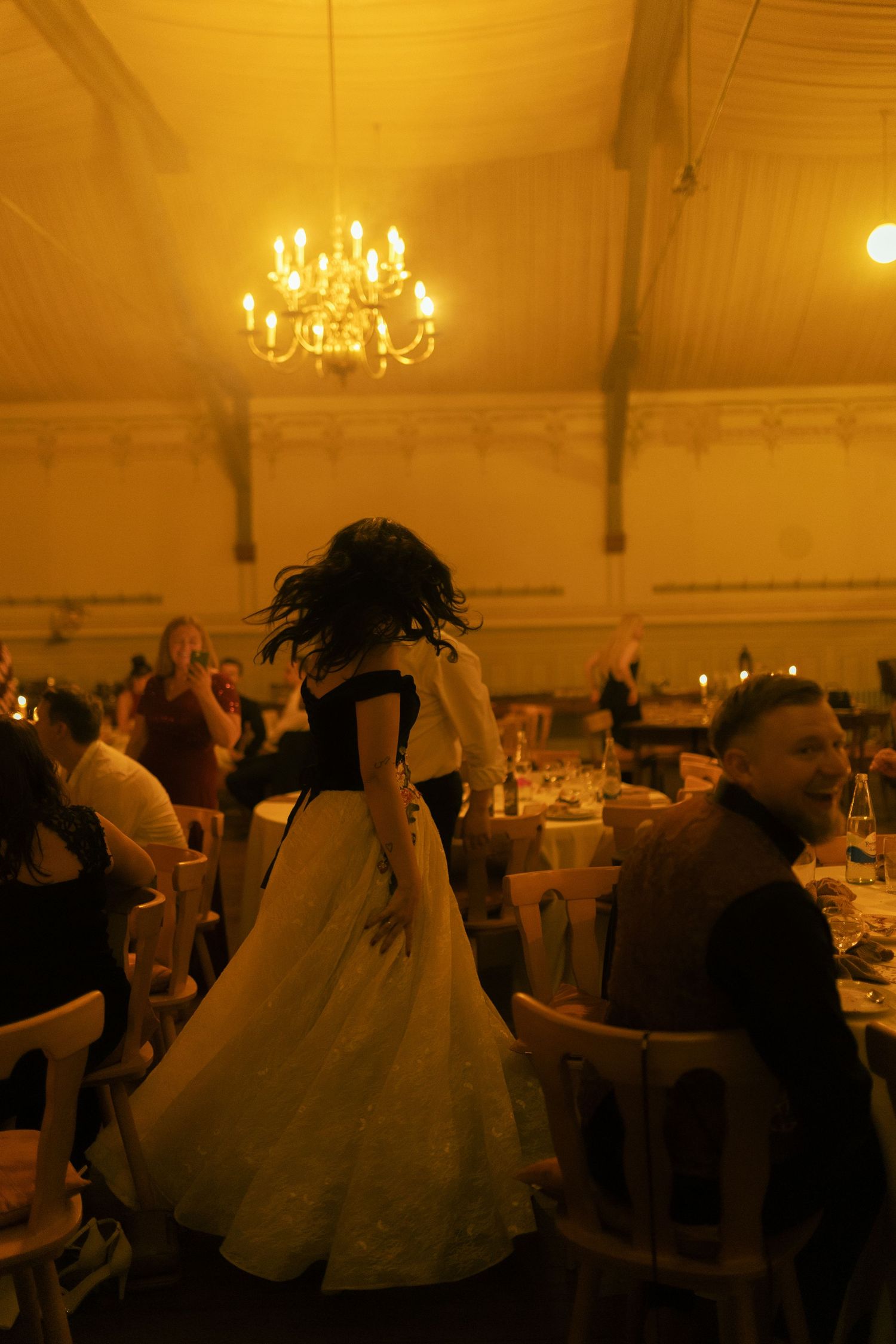 A woman in a white ballgown dances at an elegant candlelit reception hall with a chandelier glowing overhead.