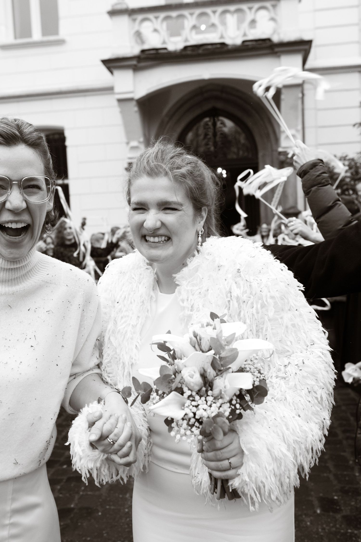 Two brides laughing joyfully outside a wedding venue, one holding a bouquet, in black and white photography.