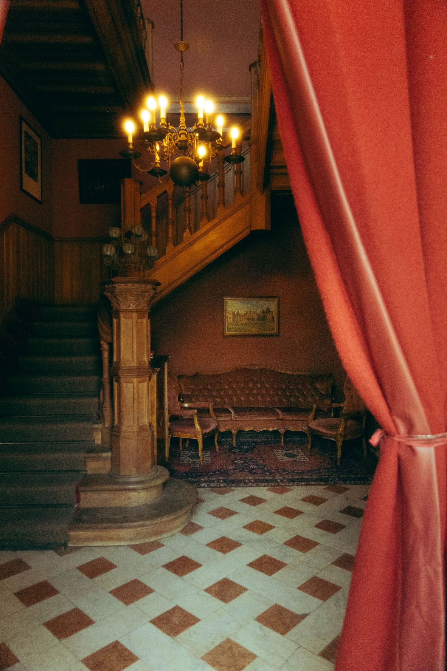 Elegant vintage foyer with red curtains, chandelier, wooden staircase, and antique sofa on a checkered tile floor.