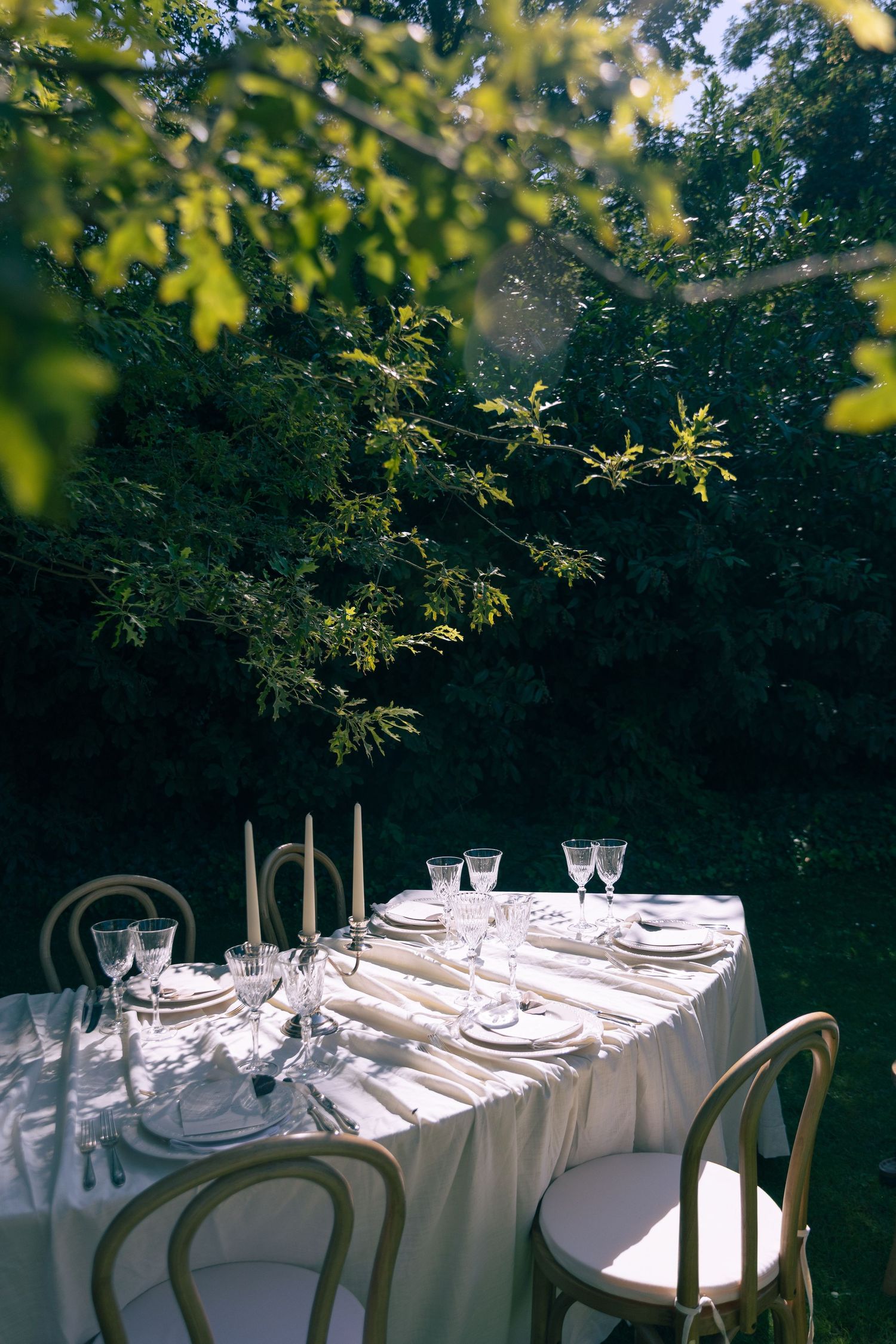 Elegant outdoor dining table set with white linen, crystal glasses, and candles surrounded by lush green trees.