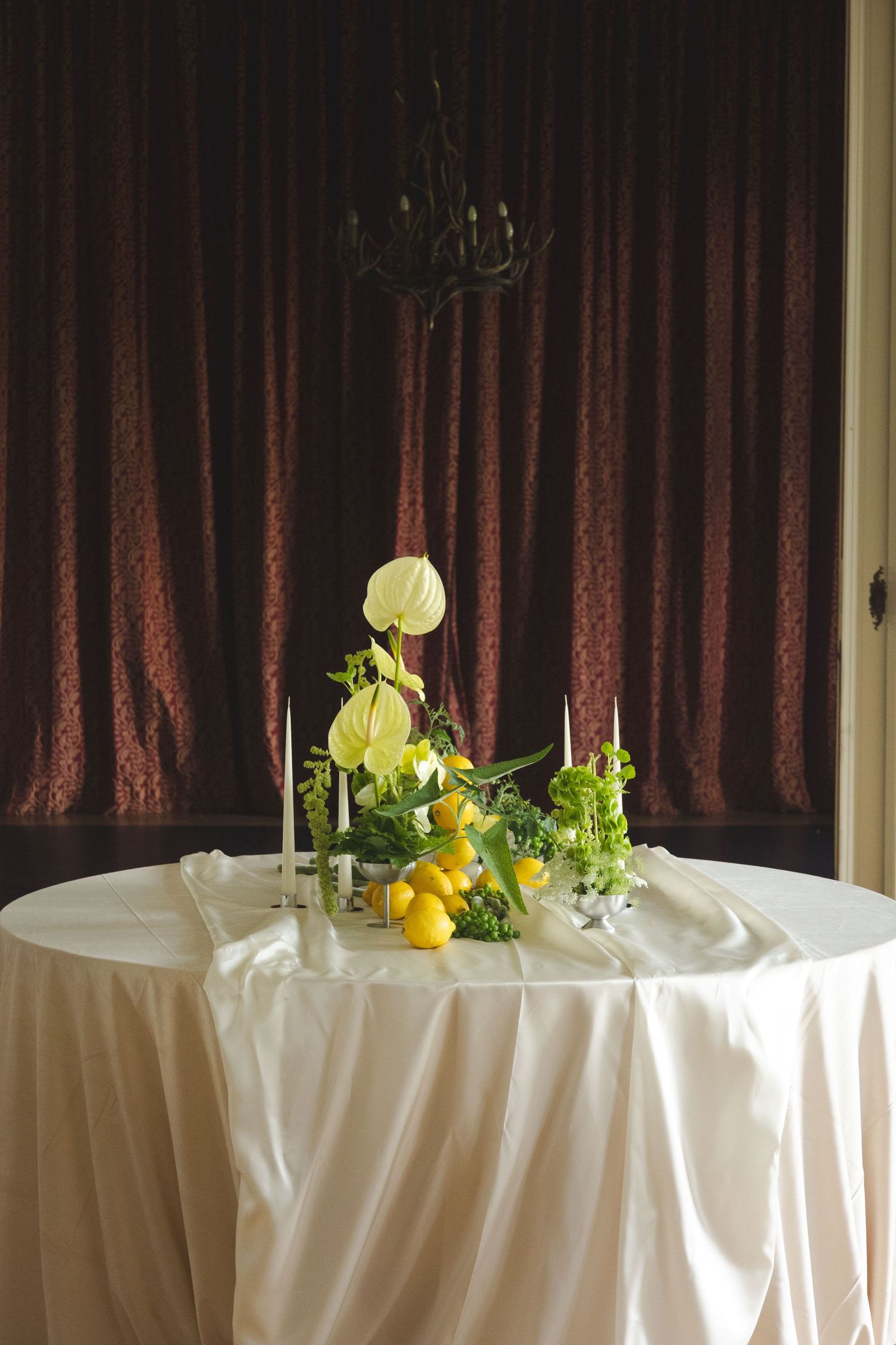 Elegant white round table with floral centerpiece featuring white anthuriums, lemons, and candles against burgundy drapes.