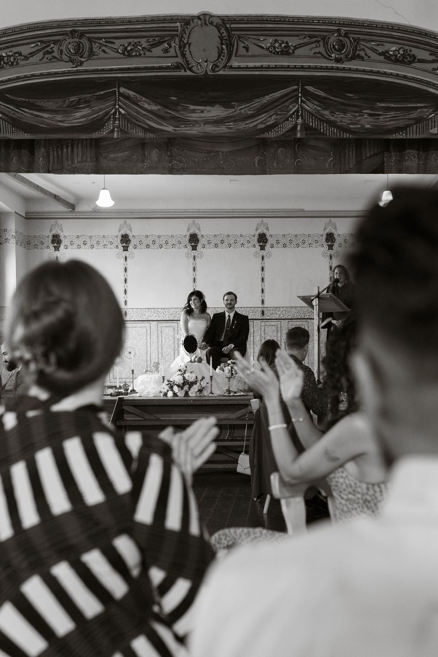 Black and white wedding ceremony photo showing couple at altar while guests applaud in elegant vintage venue.