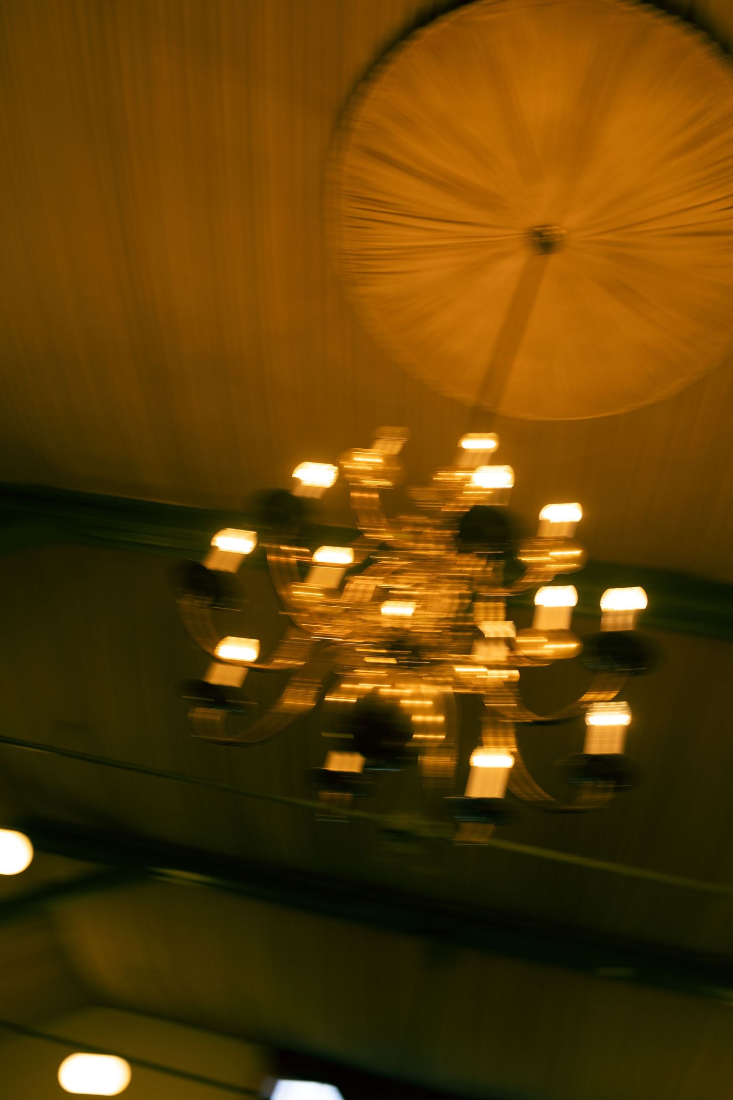 Blurry golden chandelier with glowing candle-style lights hanging from an ornate ceiling in a dimly lit room.