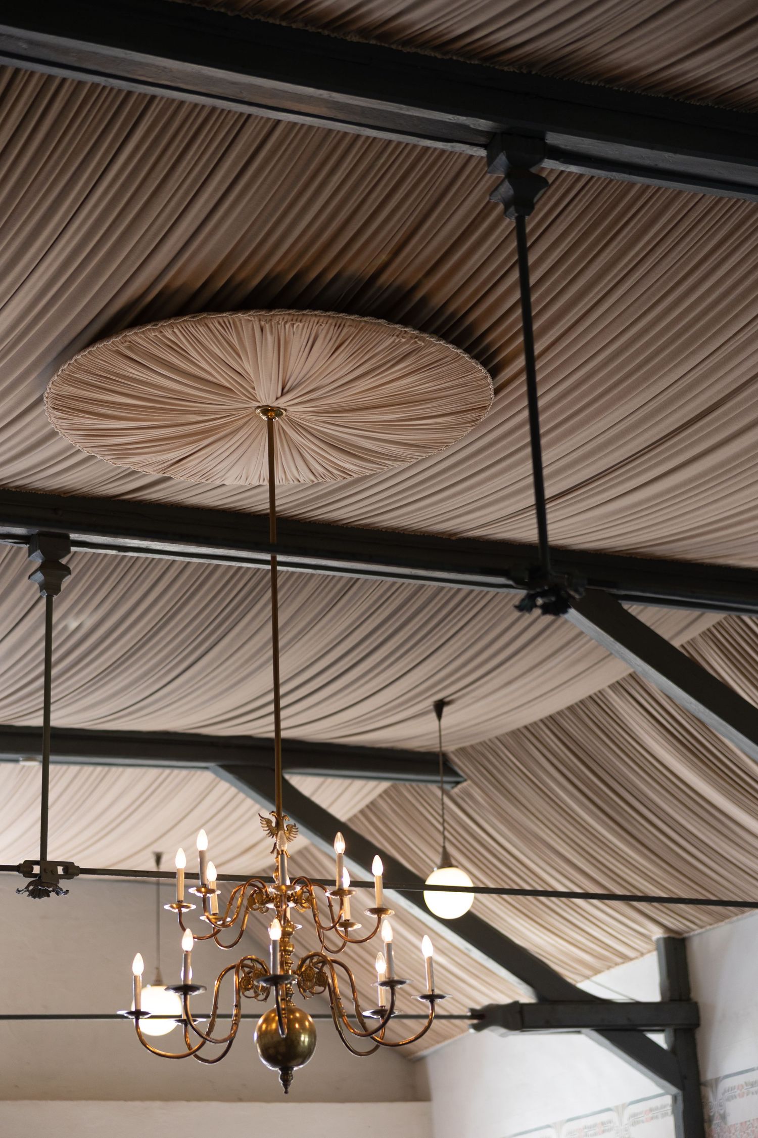 Elegant gold chandelier with candle lights hanging from a draped fabric ceiling with dark metal beams.