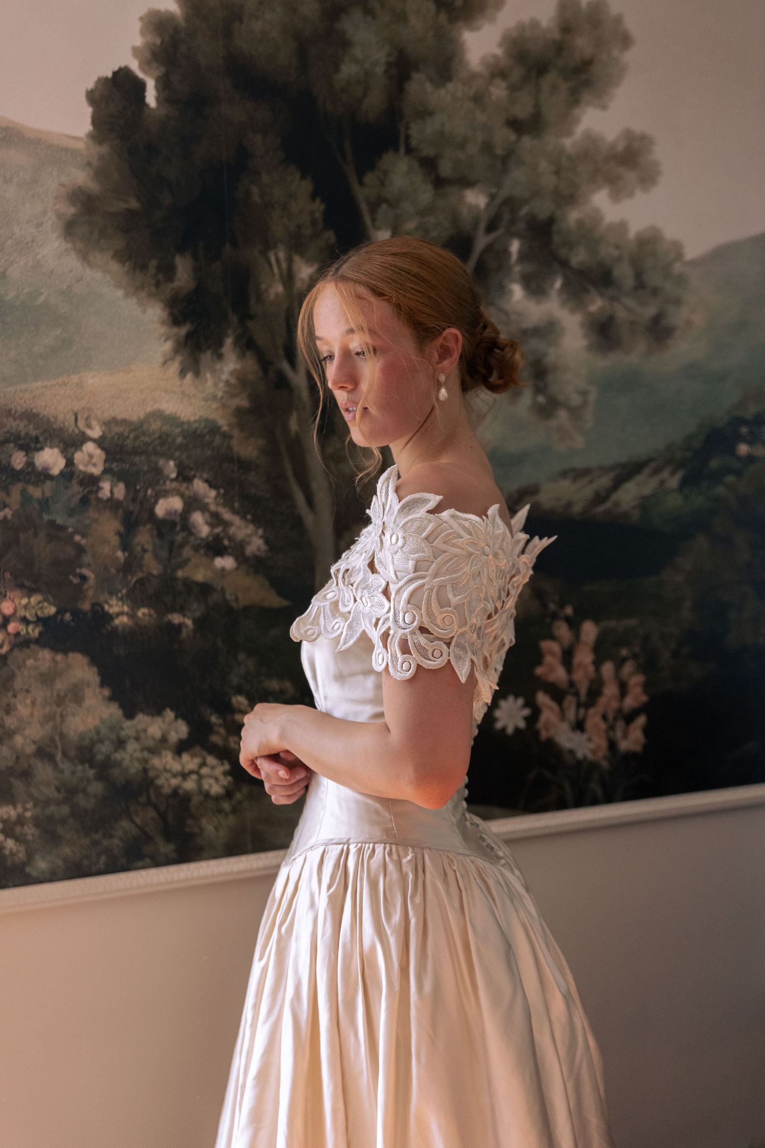 Young woman in elegant white lace off-shoulder gown stands before a classic floral landscape painting.