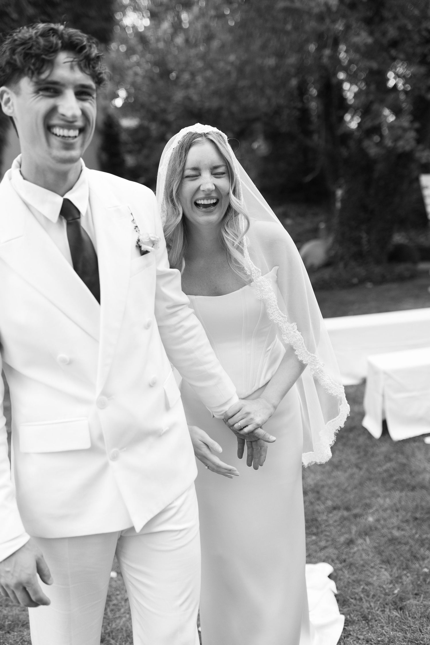 Black and white photo of a laughing bride in veil and groom in white suit celebrating outdoors at their wedding.