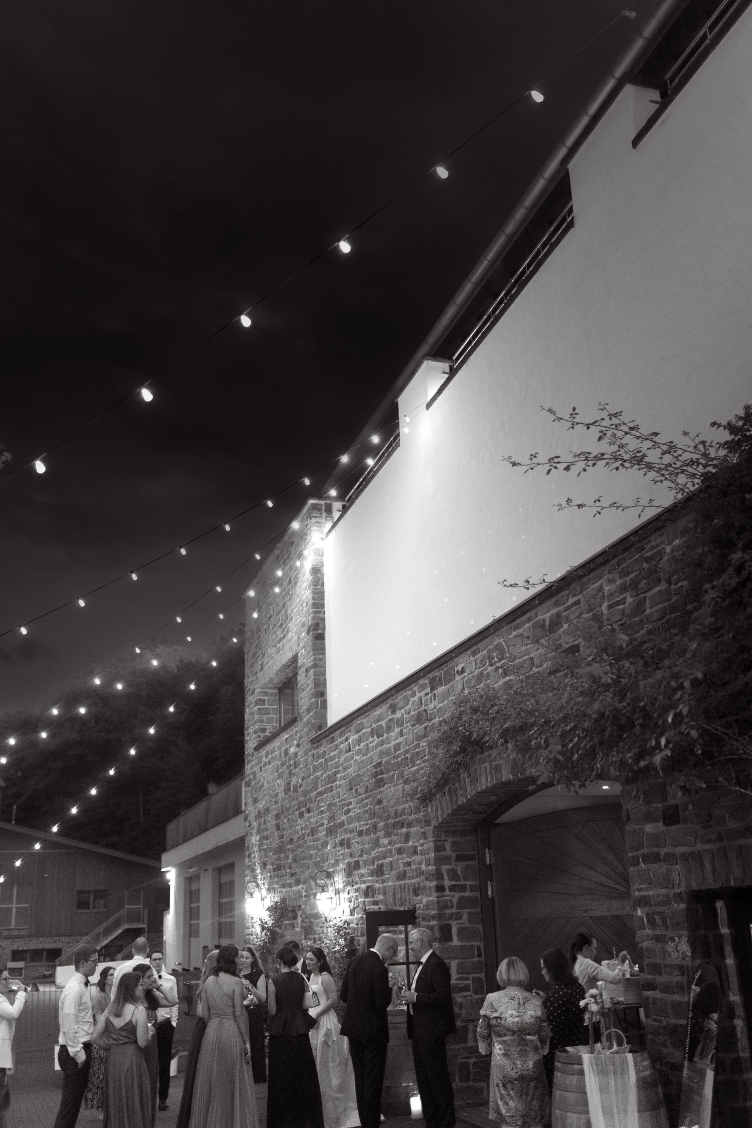 Black and white photo of wedding guests mingling outdoors at night under string lights beside a stone building.