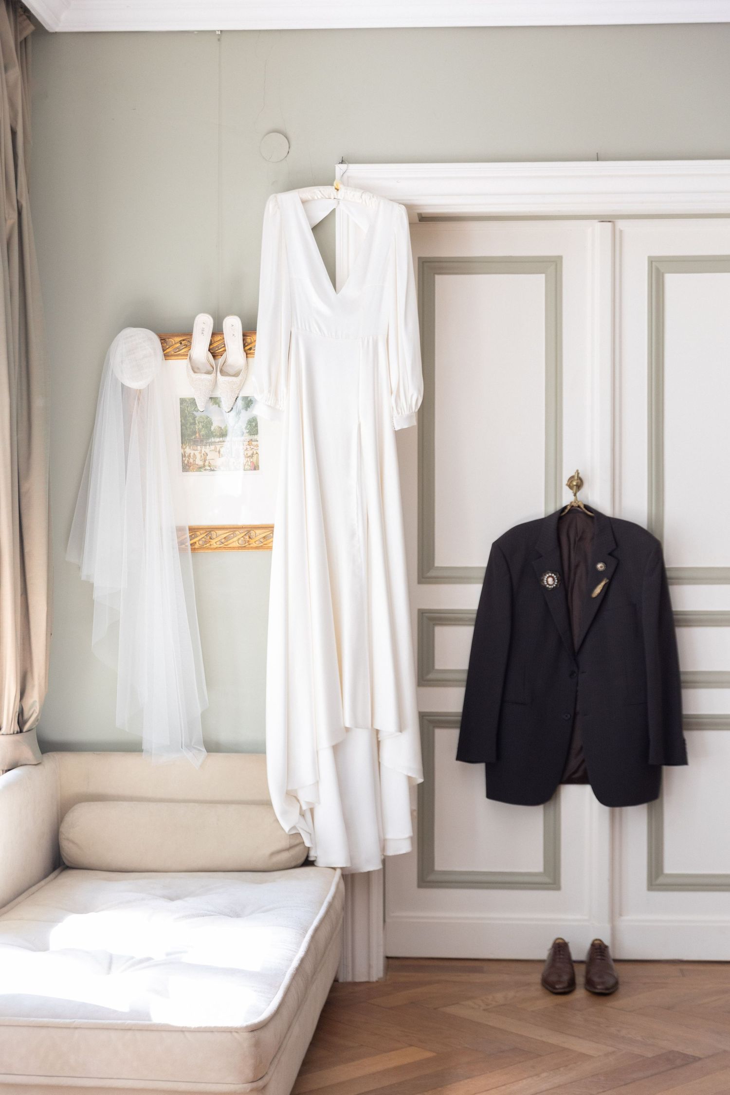 White wedding dress and dark suit jacket hanging in an elegant room with veil and bridal shoes displayed nearby.