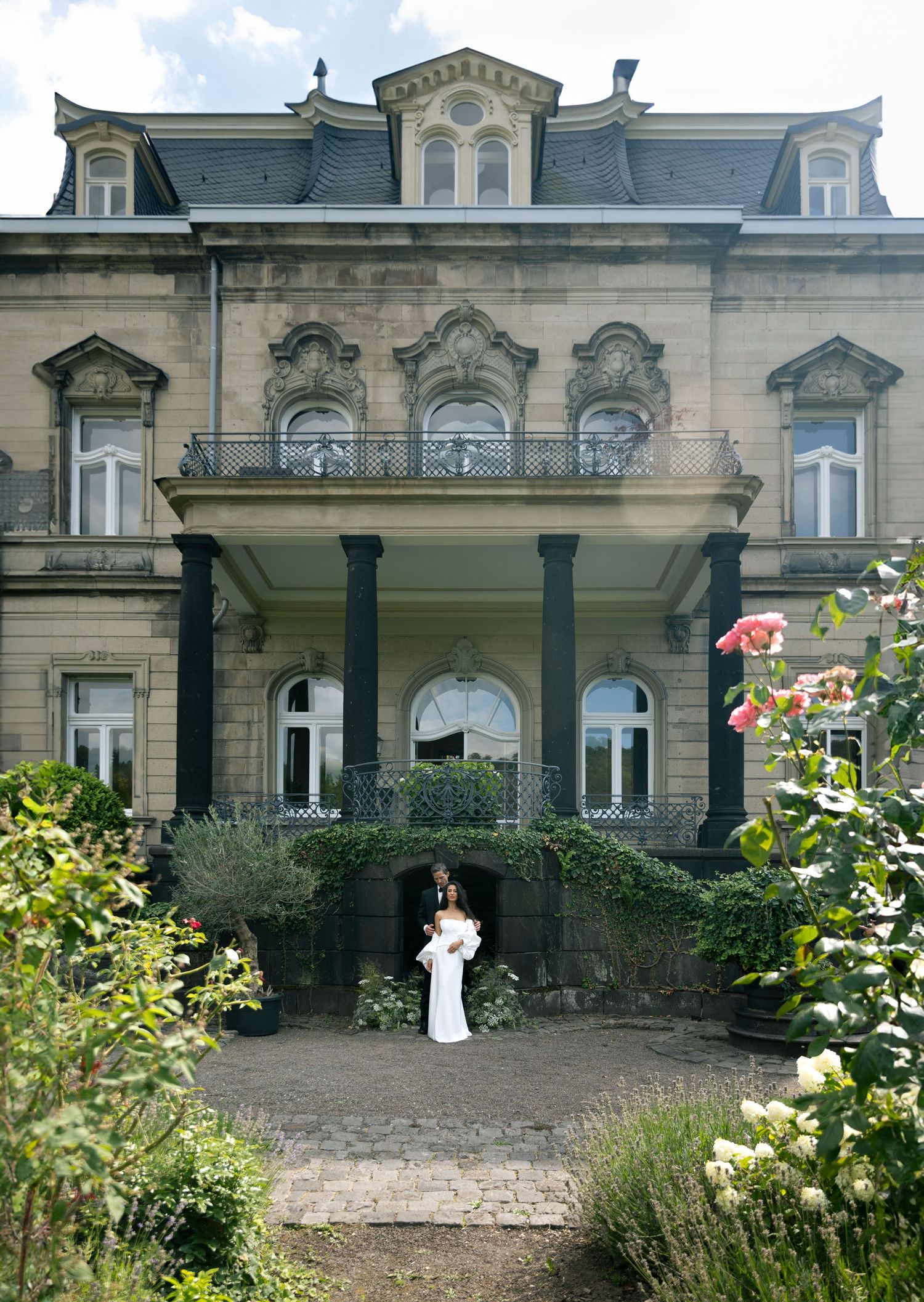Bride in white gown poses before grand historic stone villa with ornate balcony and lush garden roses.