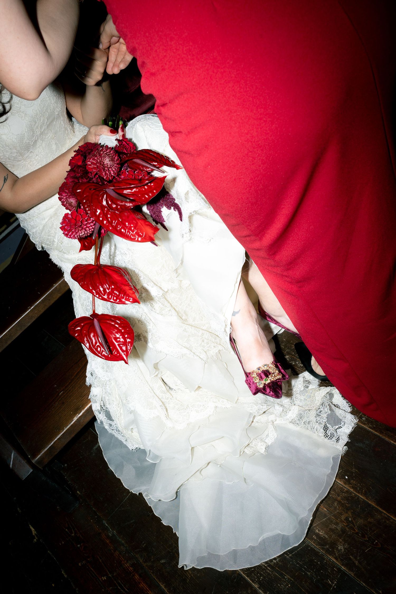 Bride in white lace gown holding red anthurium bouquet beside person in red dress on dark wood floor.