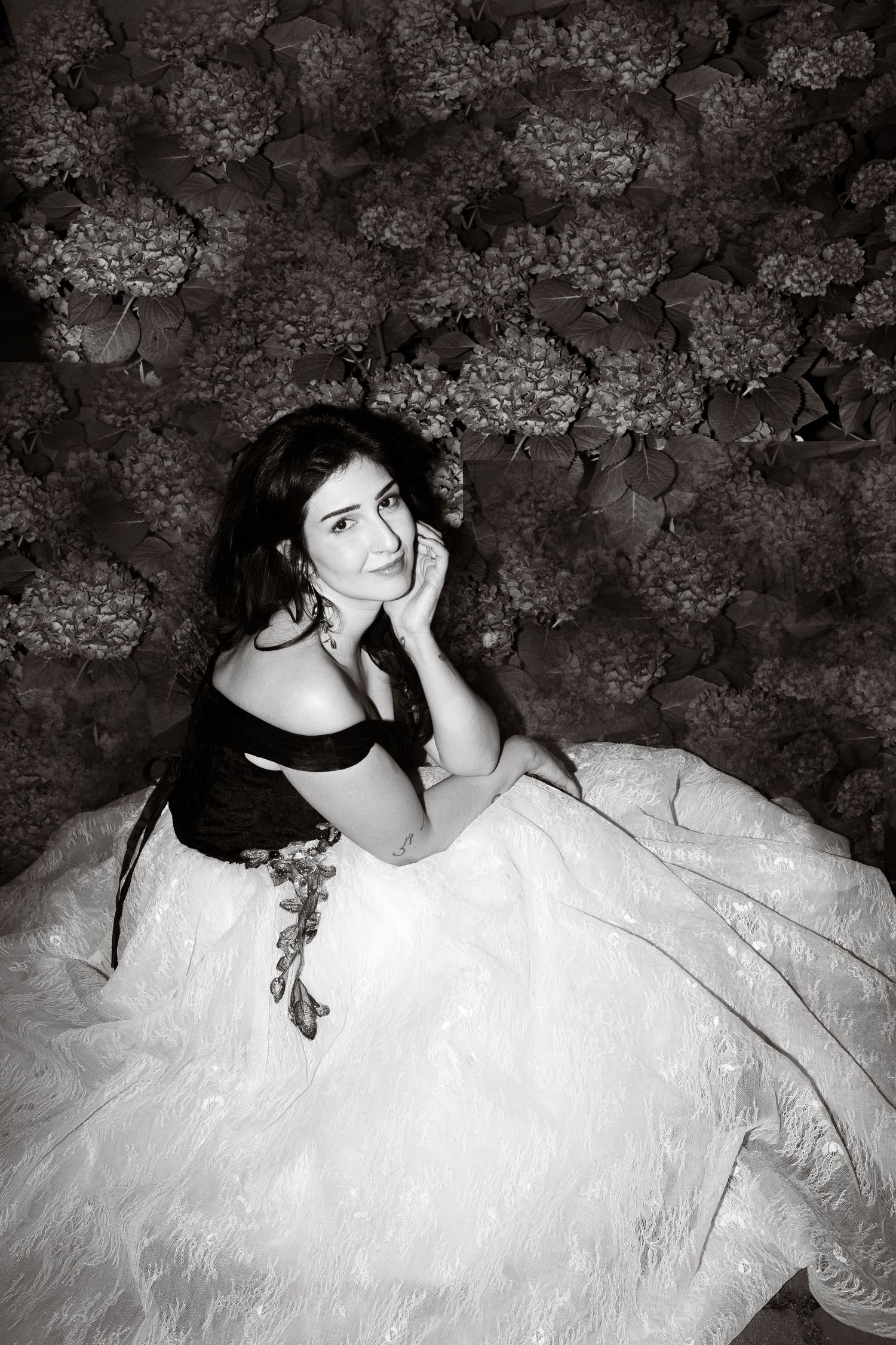 Black and white portrait of a woman in an elegant ball gown seated against a floral hydrangea backdrop.