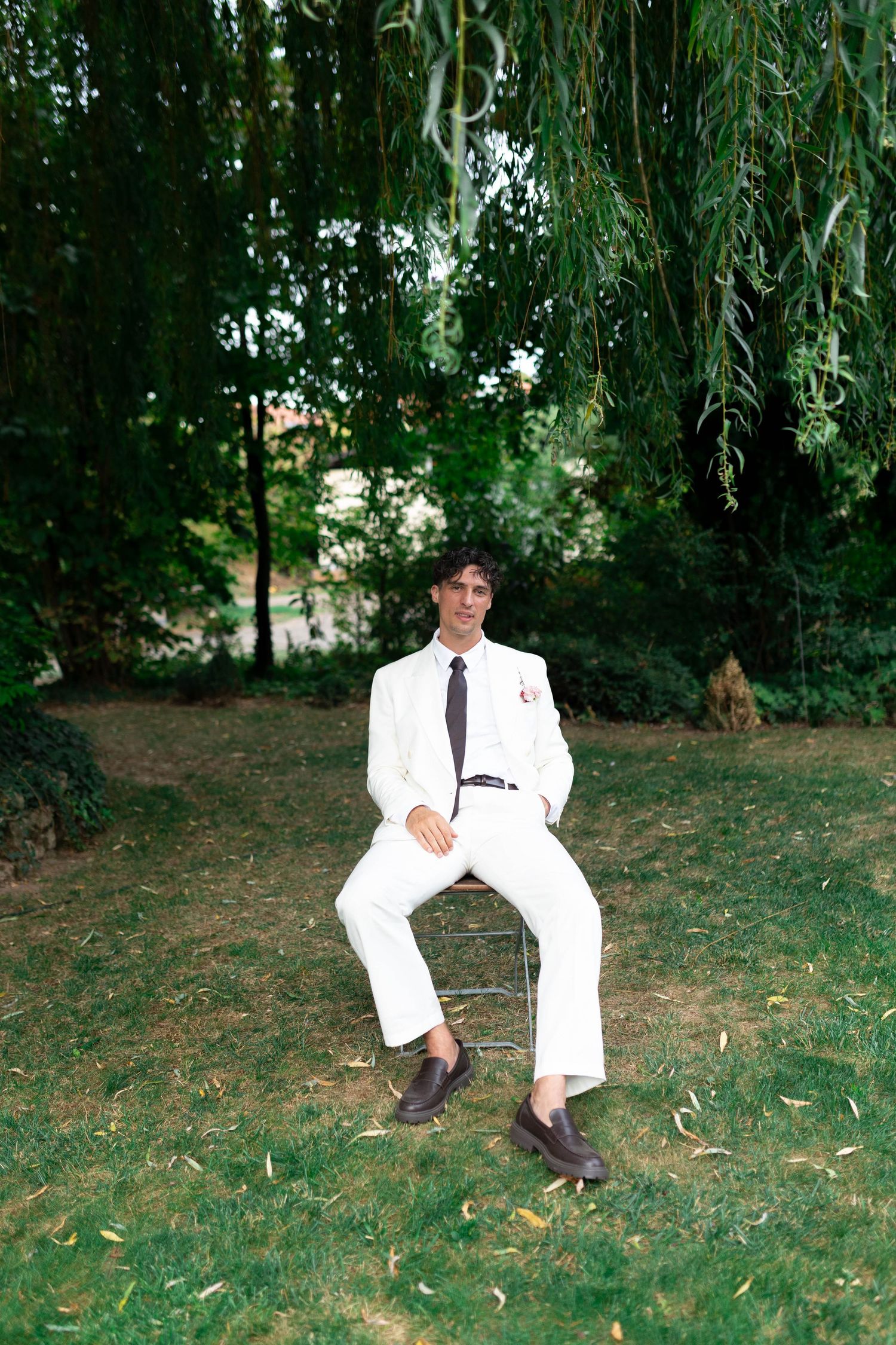 A young man in a white suit and dark tie sits on a chair under a weeping willow tree in a lush green garden.