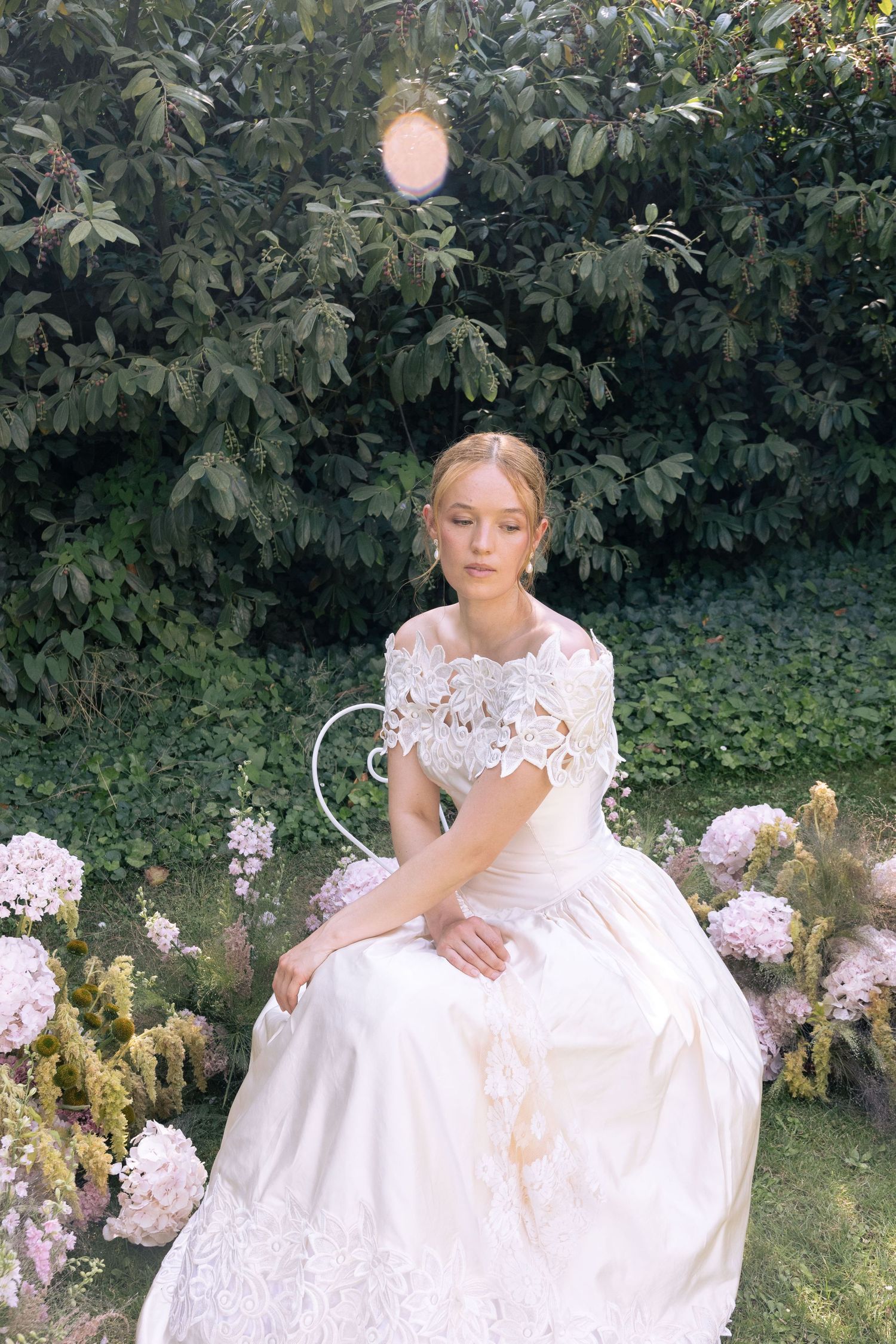 A bride in a white lace gown sits gracefully among blooming hydrangeas in a lush green garden.