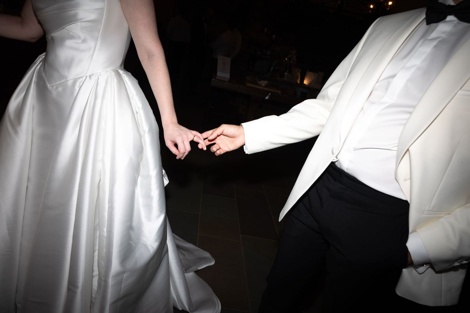 Bride and groom holding hands while dancing at wedding reception, wearing white gown and white tuxedo jacket.