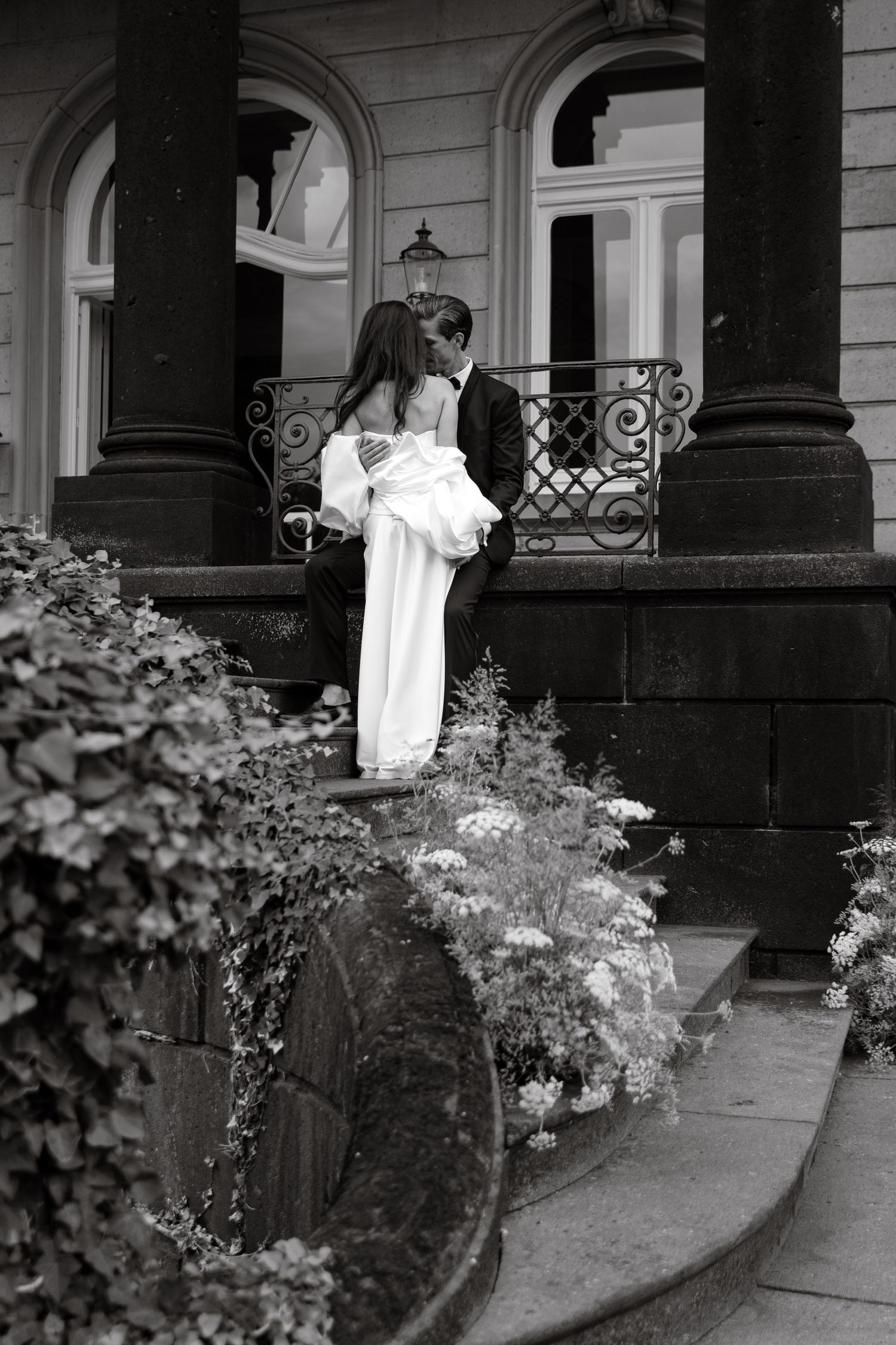 Black and white photo of a couple kissing on elegant stone steps of a historic building surrounded by lush garden.