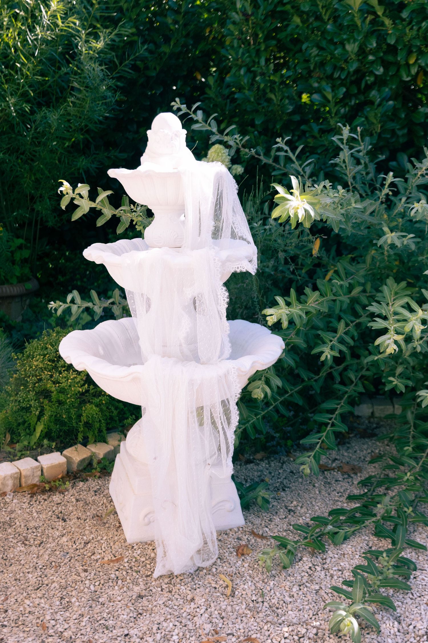 White tiered garden fountain draped with sheer tulle fabric, surrounded by lush green shrubs and gravel landscaping.