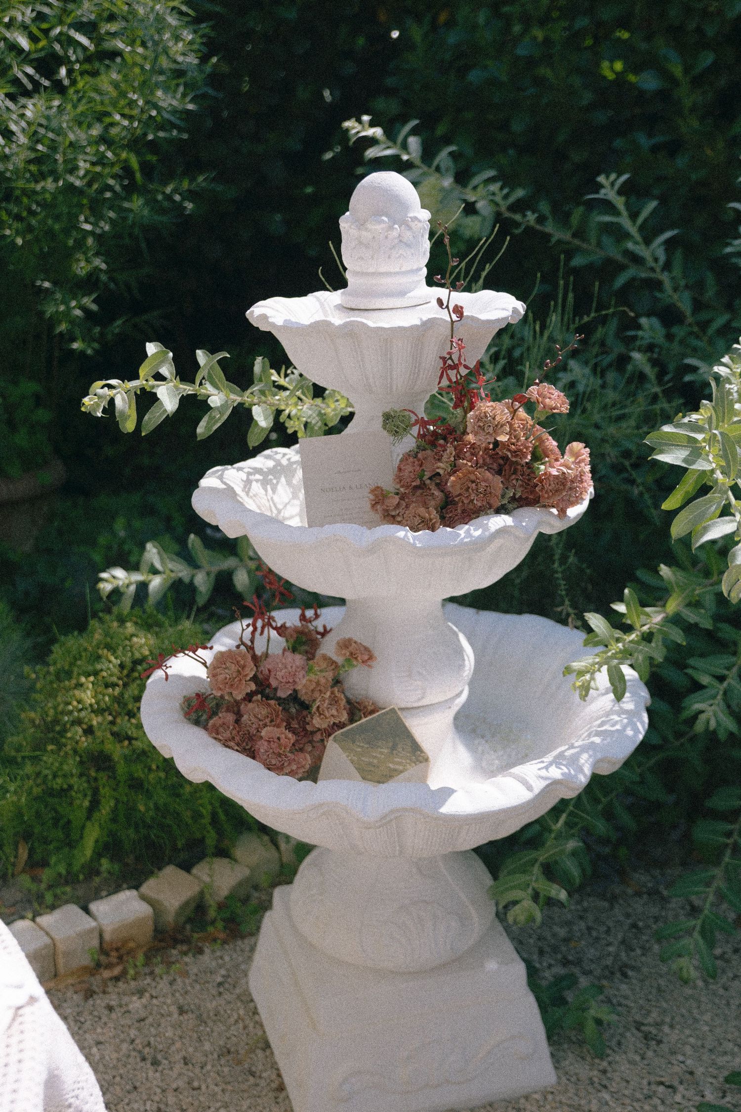 A white three-tier garden fountain decorated with dried pink flowers, surrounded by lush green shrubs.